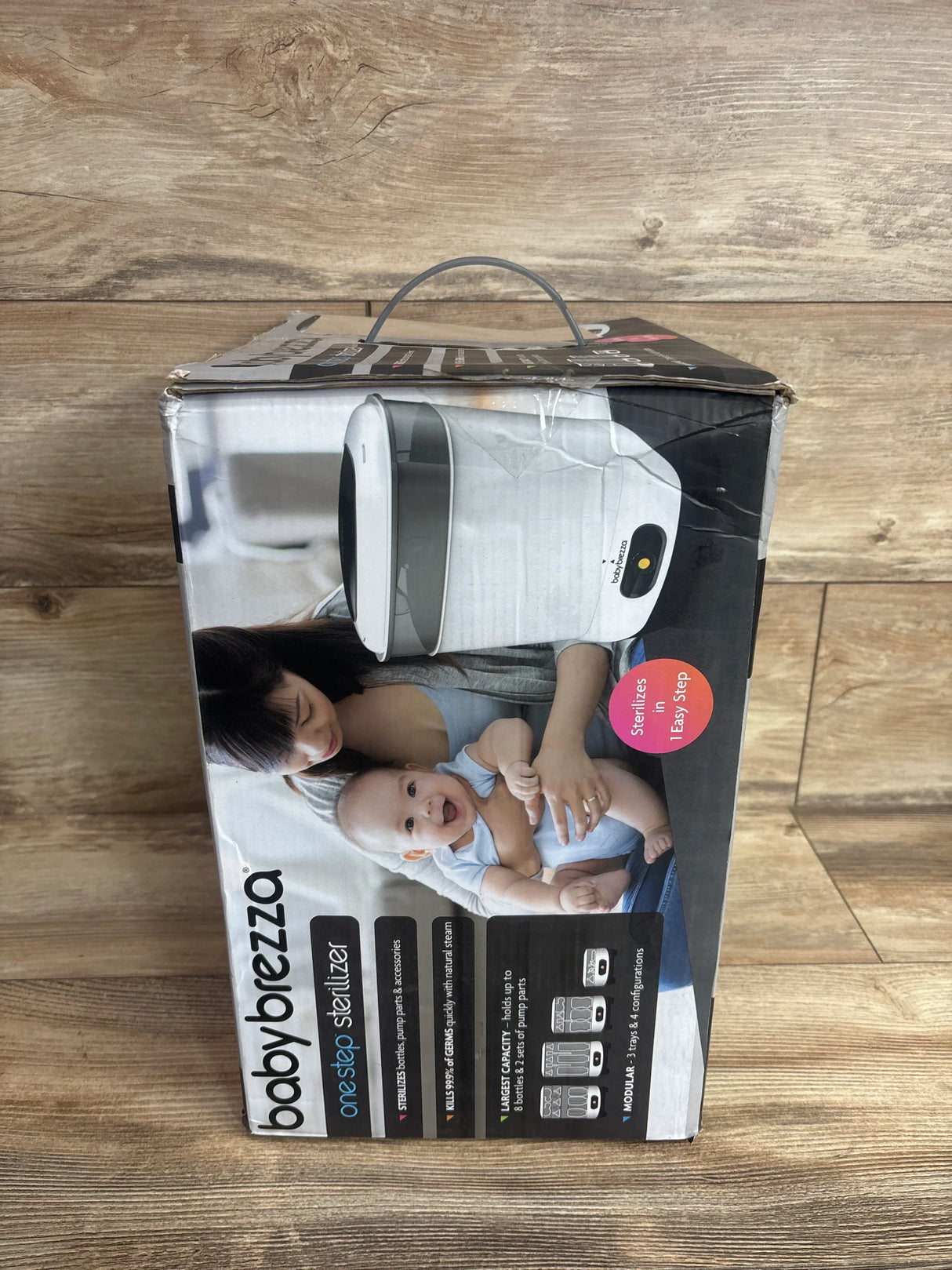 Angled front view of Baby Brezza One Step Sterilizer box showing product photo and the overlaid text Sterilizes in 1 Easy Step on a wooden floor background