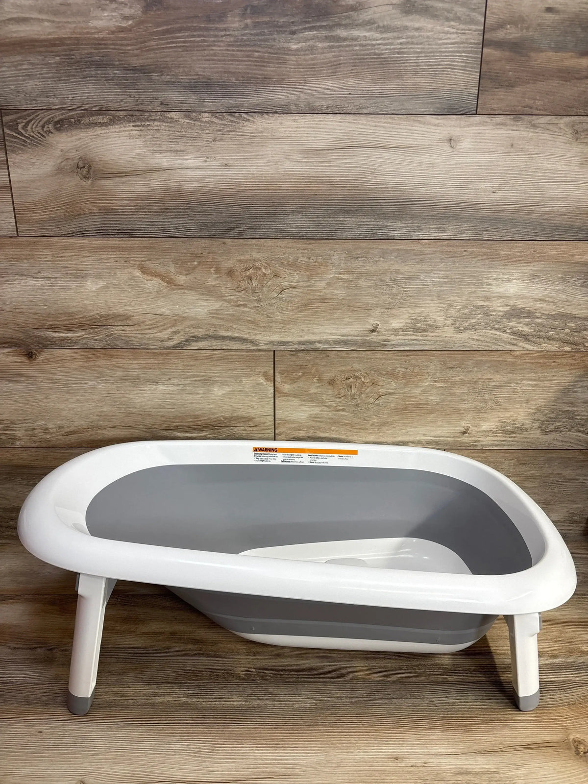 Three-quarter view of OXO Tot Splash and Store Bathtub in grey showing foldable legs and white interior on wood floor