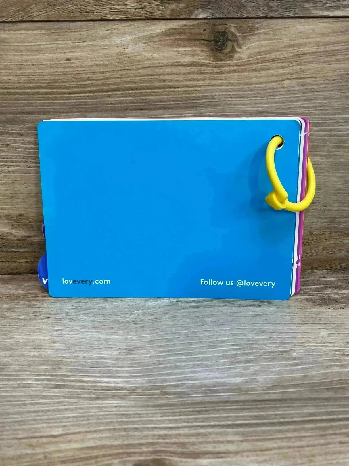 Back view of Lovevery Babbler play guide (13-15m) showing blue back, yellow ring binder, and colorful page edges on wood