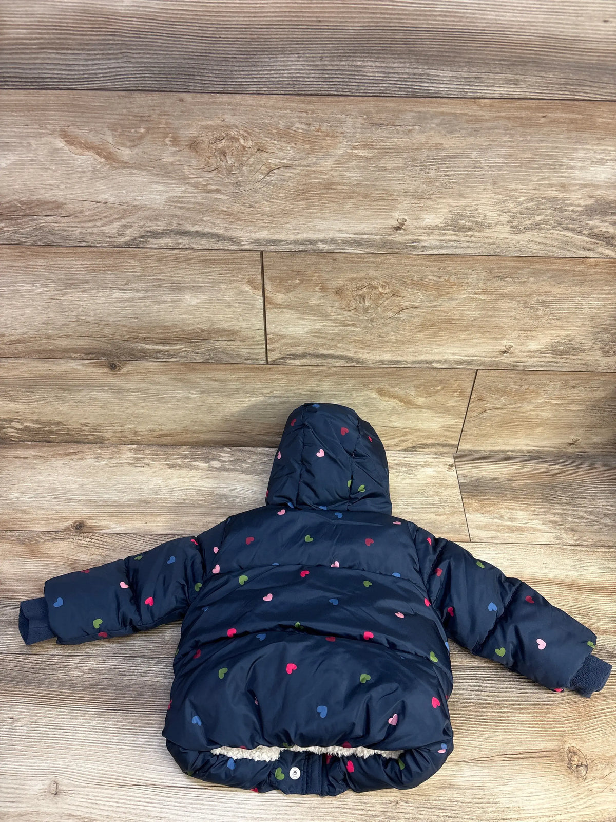 Back view of Baby Gap navy puffer jacket with hood, multicolor heart print and sherpa-lined hem on wood background