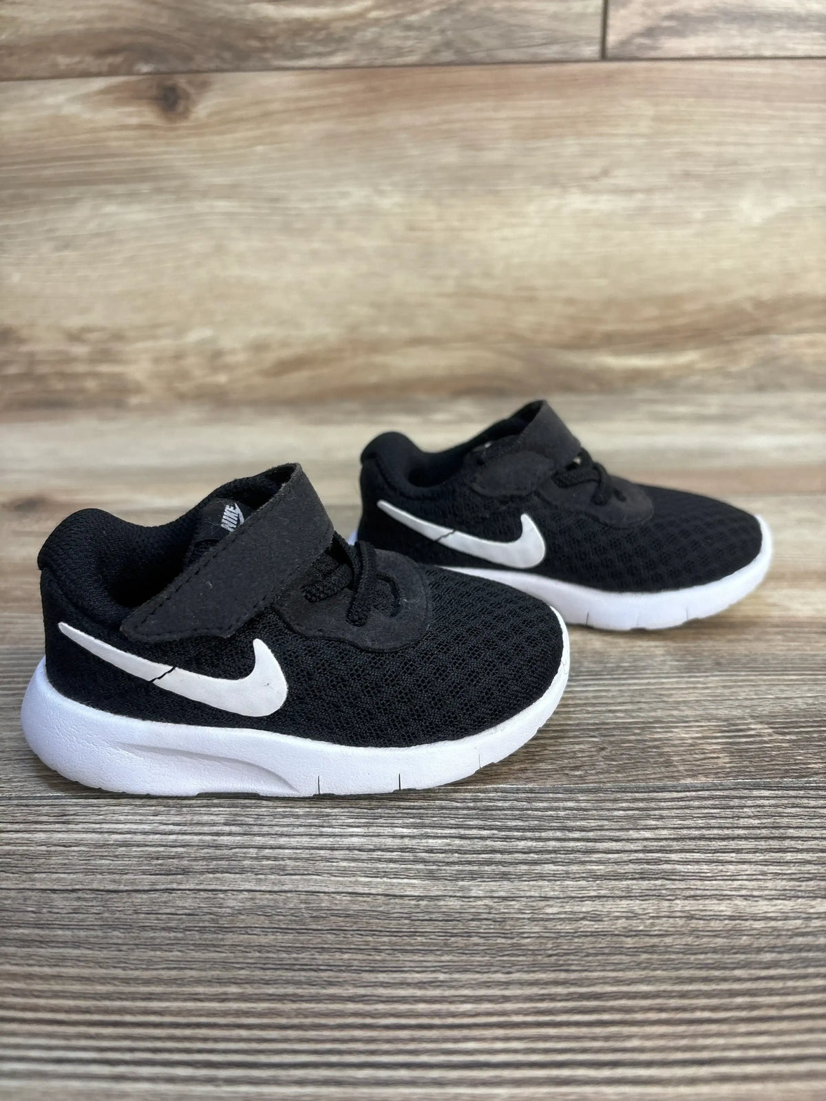 Side view of Nike Tanjun TDV kids sneakers with black mesh upper, white Swoosh, Velcro strap and white sole on wood background