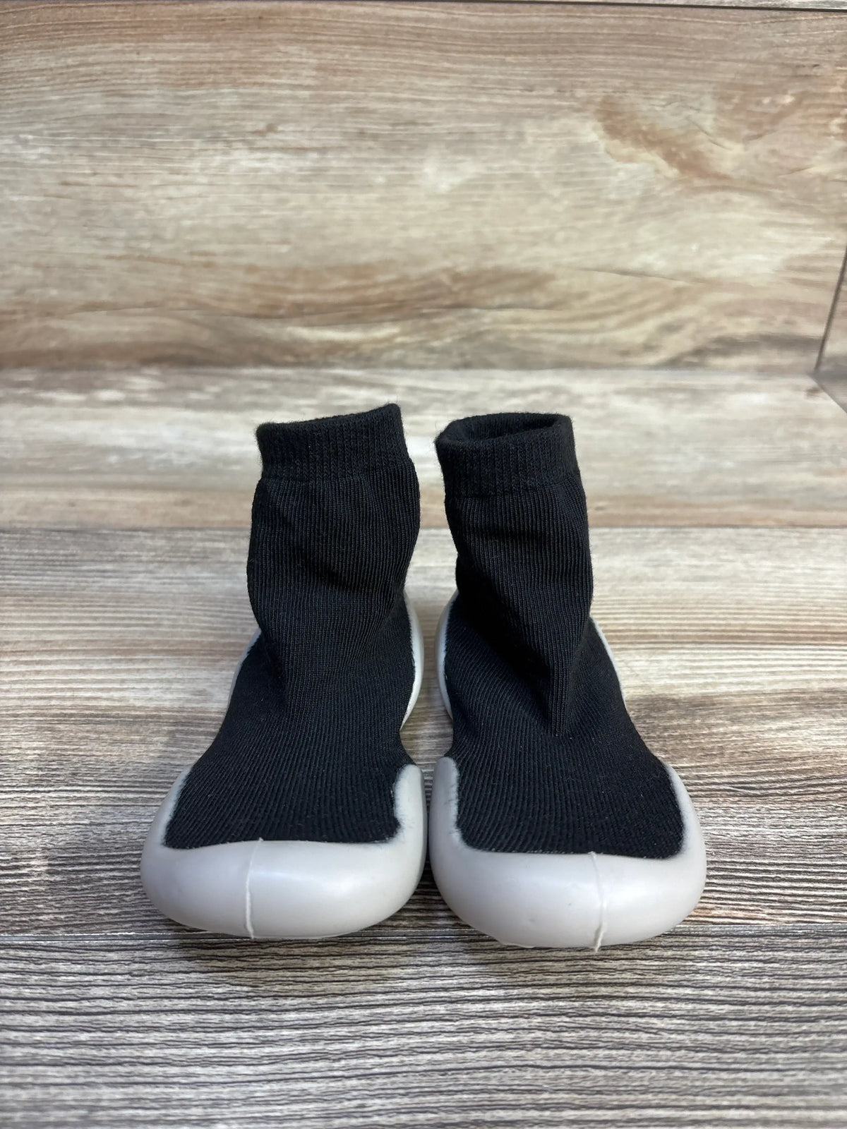 Front view of black baby sock shoes in size 6/7c showcasing sock-like uppers and white rubber soles on a wood surface