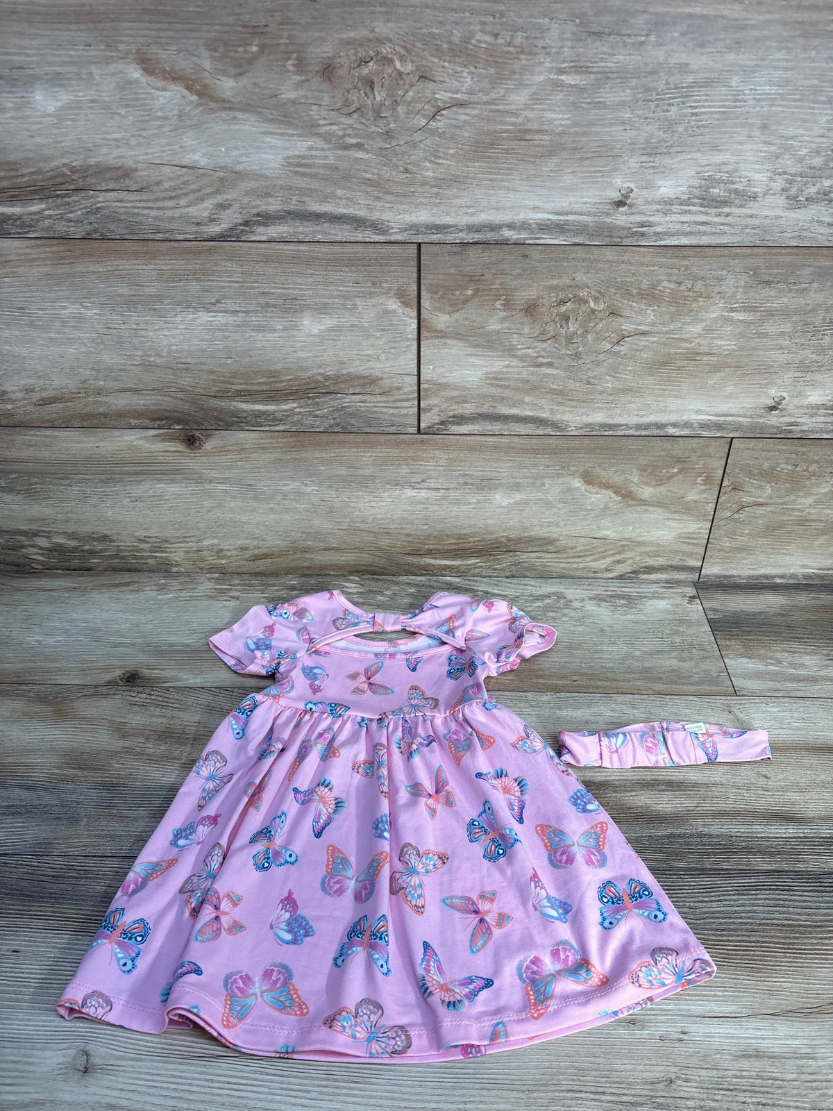 Flat lay of Picapino pink butterfly dress and matching headband set showcasing colorful butterfly print on a wood-look background