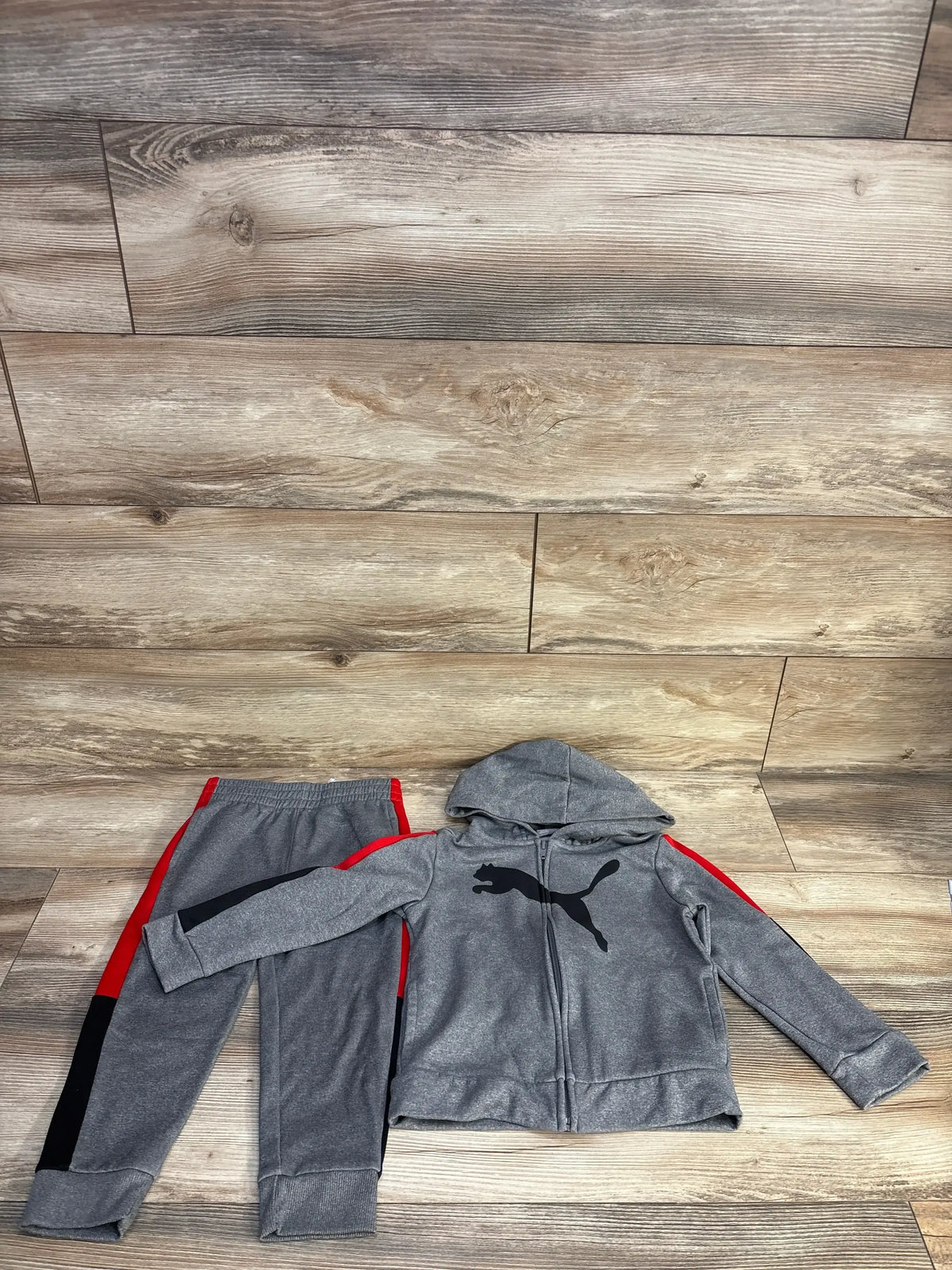 Flat-lay of grey Puma 2-piece full-zip toddler tracksuit (4T) showcasing hooded jacket with black Puma logo and red side stripes on wood floor