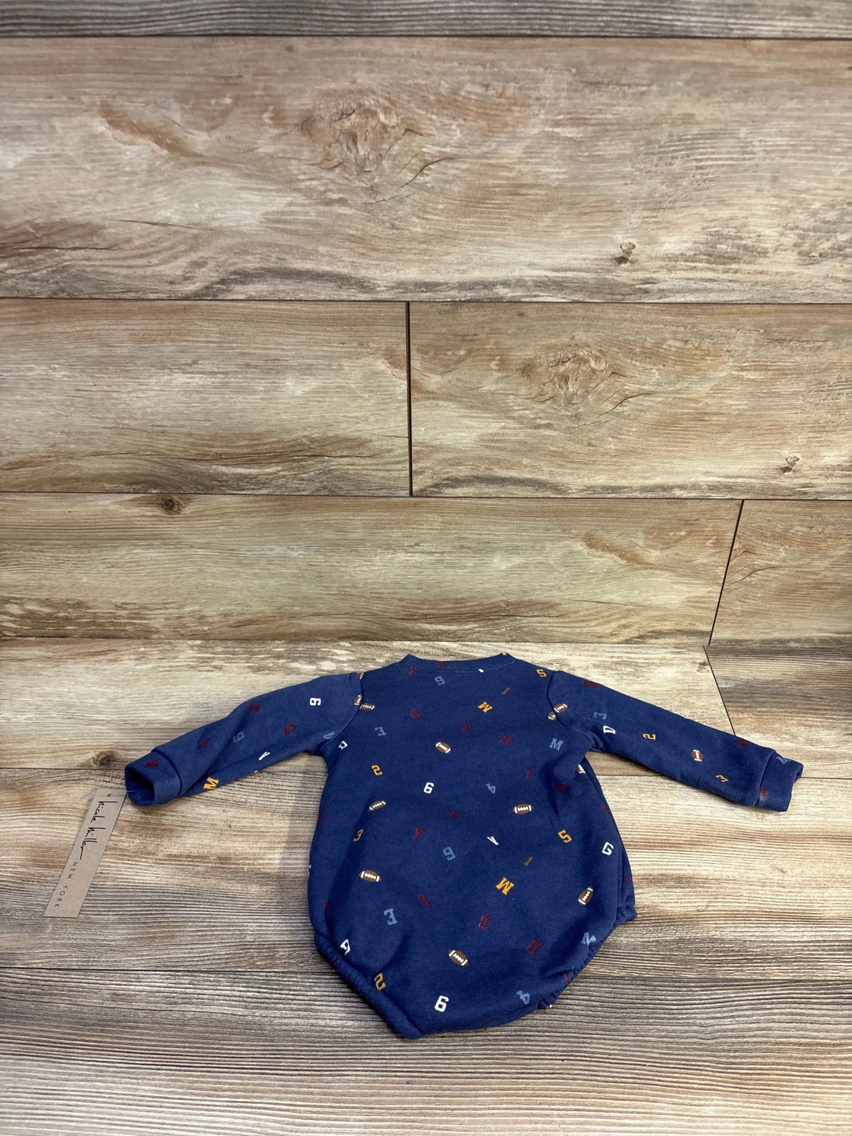 Flat lay back view of Nicole Miller football henley bubble romper showcasing navy blue fabric with small football and number print on wooden background