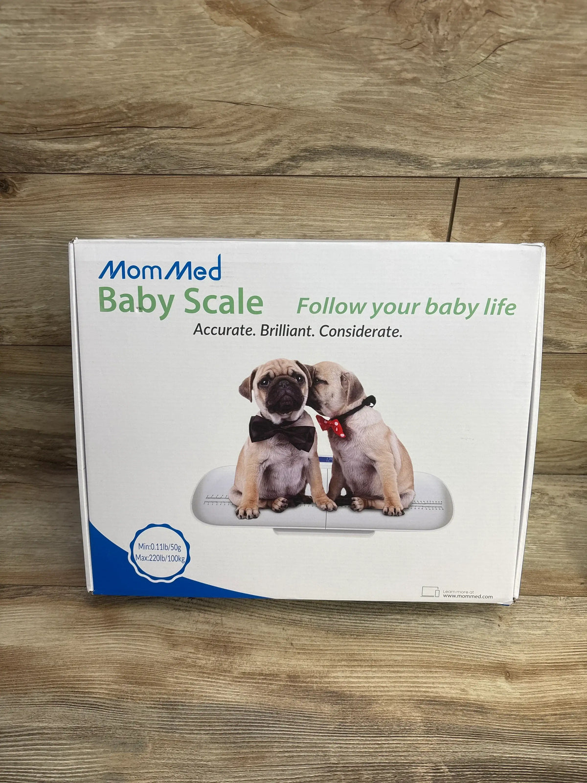 MomMed baby scale box with two puppies wearing bowties, displayed on wood floor