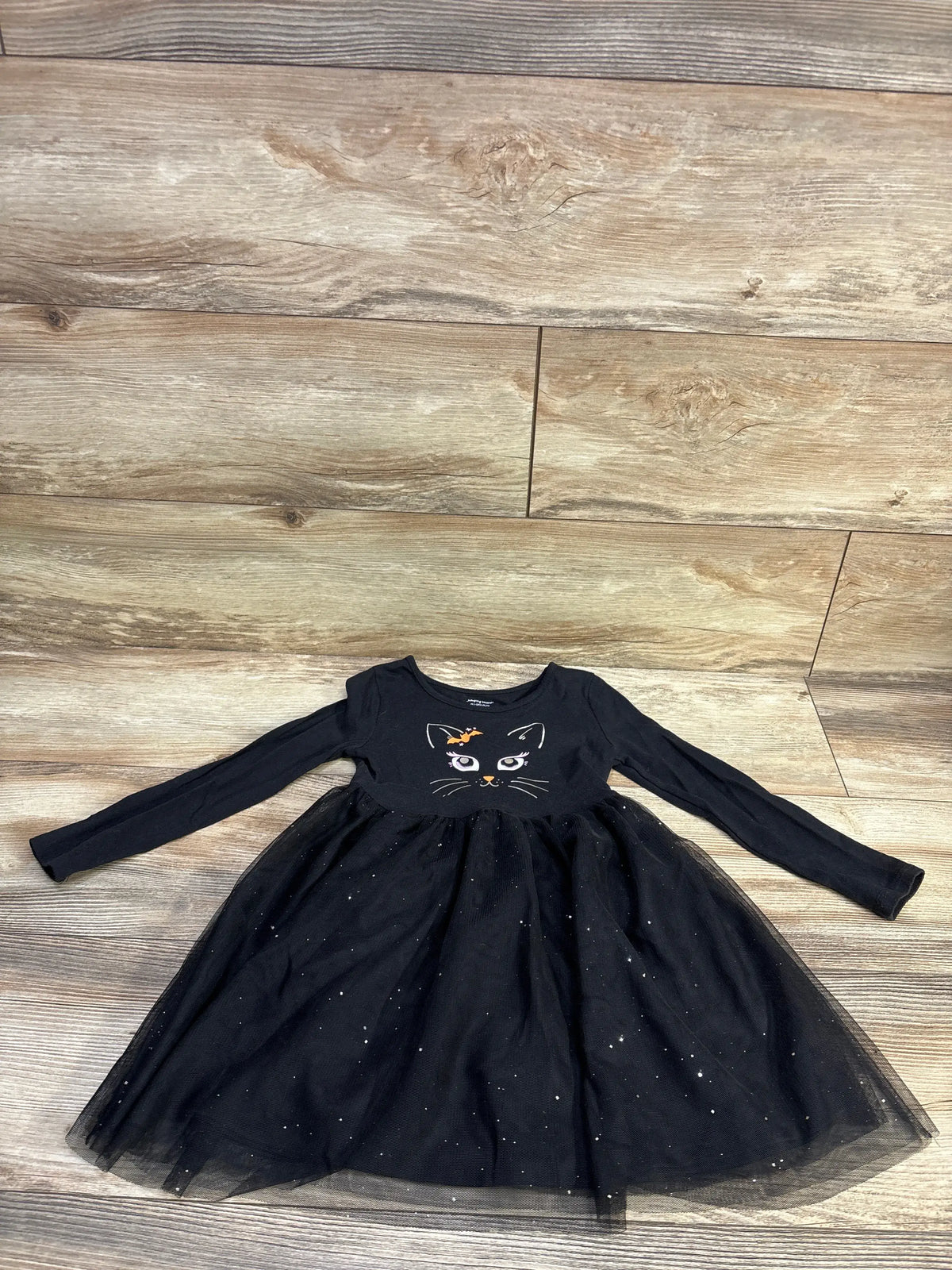 Flat lay of size 4T black Halloween cat long-sleeve dress showing cat face graphic and glitter tulle skirt on wood floor