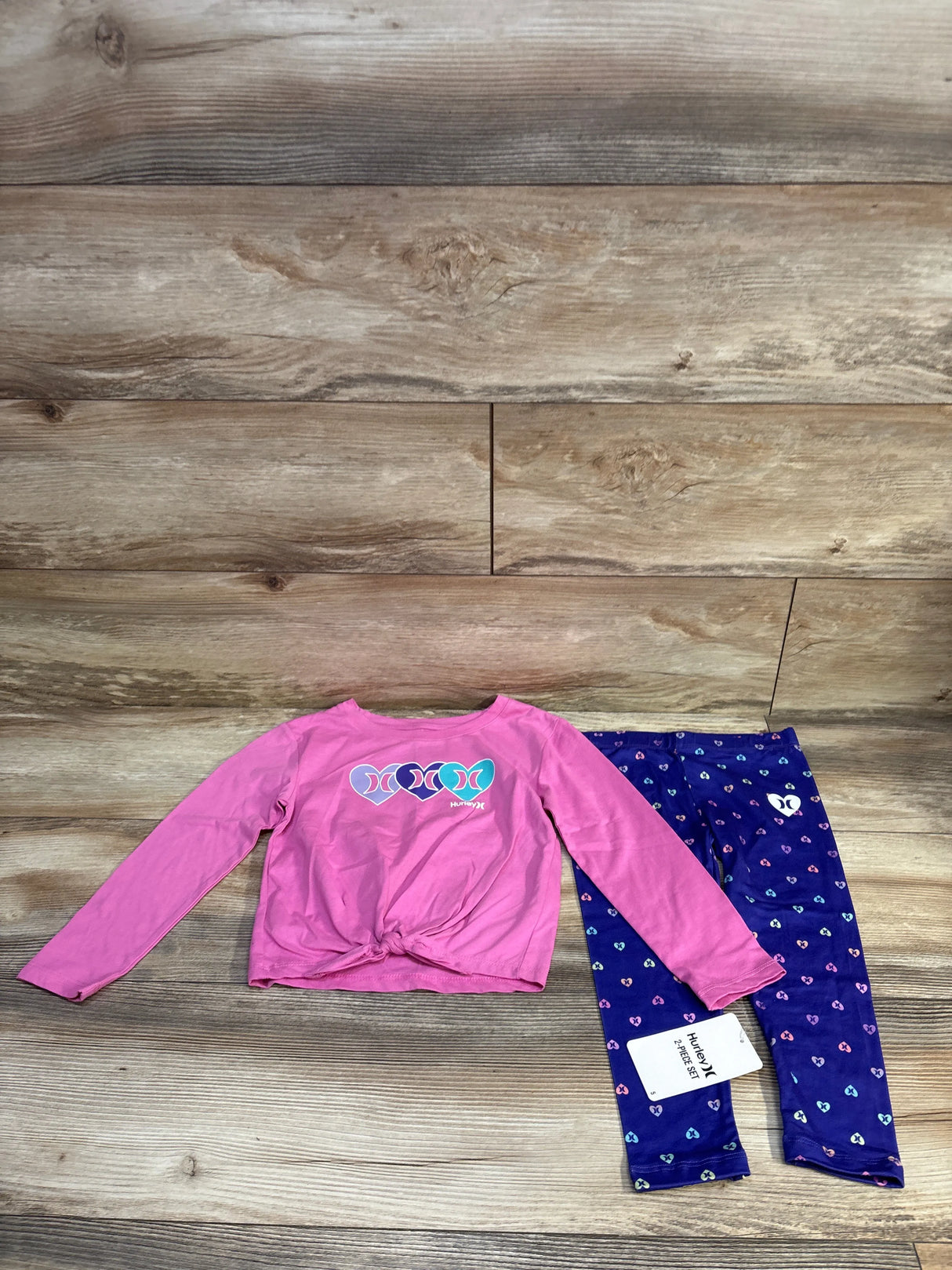 Top-down of Hurley pink long-sleeve knot-front shirt and navy heart-print leggings showing knotted hem, logo and size tag on wood background
