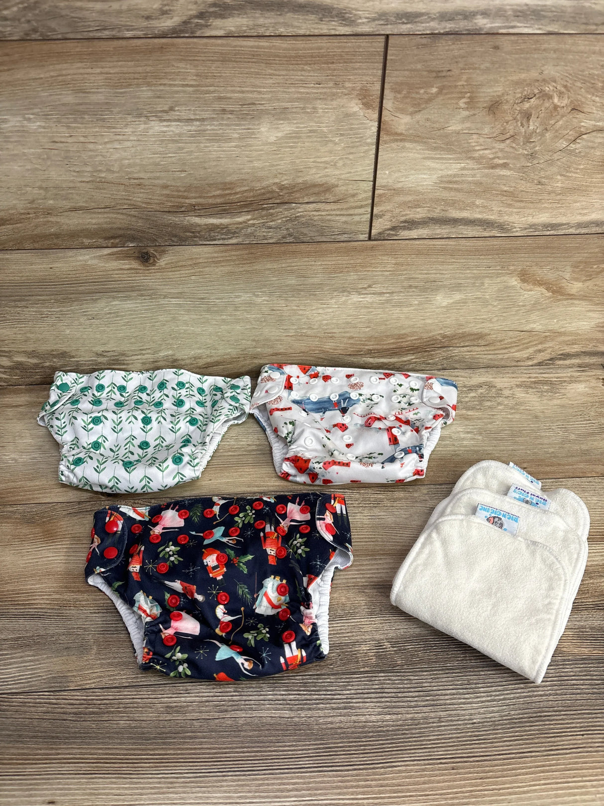 Top-down of Rig ‘N Gigs pocket cloth diapers showing three printed covers (green, fox, navy) and two inserts on wood floor