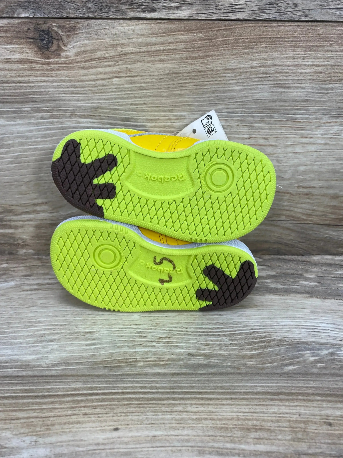 Soles-up view of toddler Minions x Club C 85 sneakers with lime-green soles and brown Minion footprint on wood background