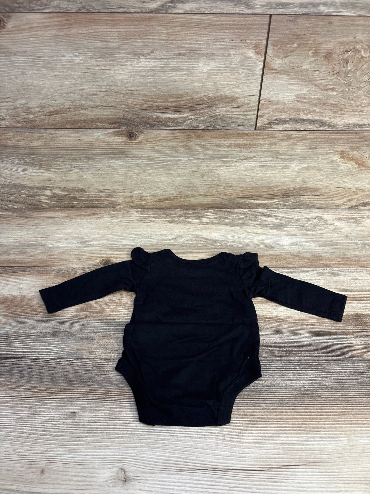 Black long-sleeve baby onesie with ruffle shoulder details laid flat on wooden floor