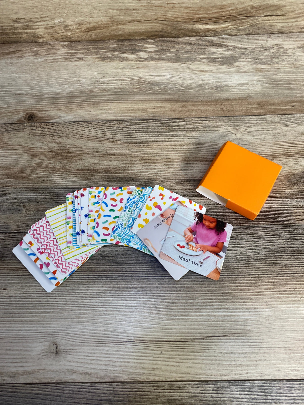 Colorful educational flashcards spread on wooden table with open orange box, meal time card visible