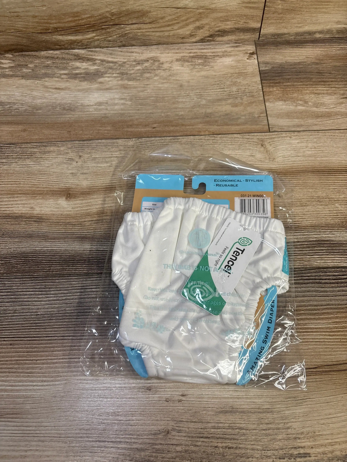 Top-down view of Charlie Banana white swim diaper size L (18-36 mo) showing elastic waistband and blue trim in packaging on wood floor