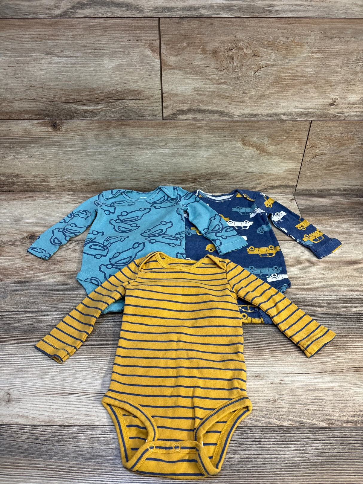 Front of three Carter's long-sleeve 9M bodysuits: mustard stripe, light blue print, navy with yellow vehicle print on wood