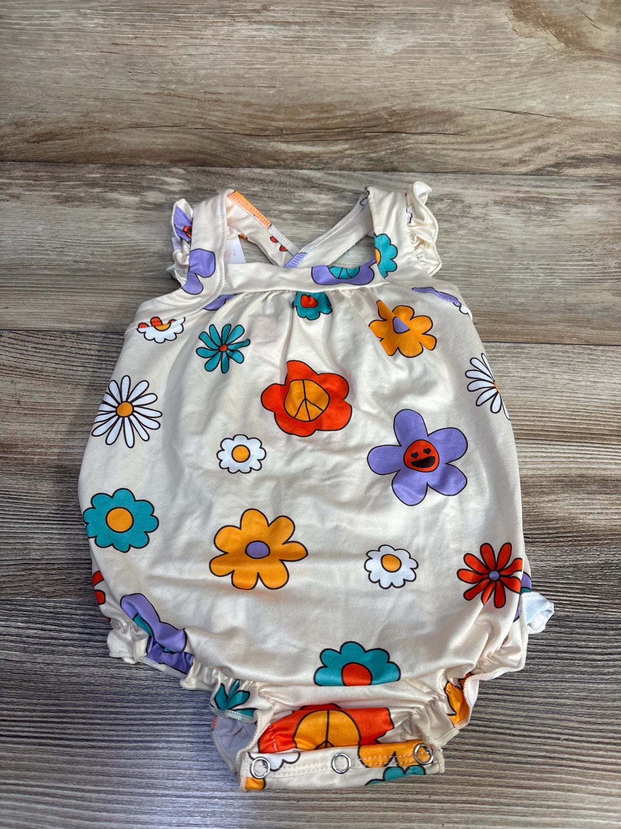 Top-down of beige Mila & Rose floral crossback romper with colorful flowers, ruffle straps, and snap crotch on light wood