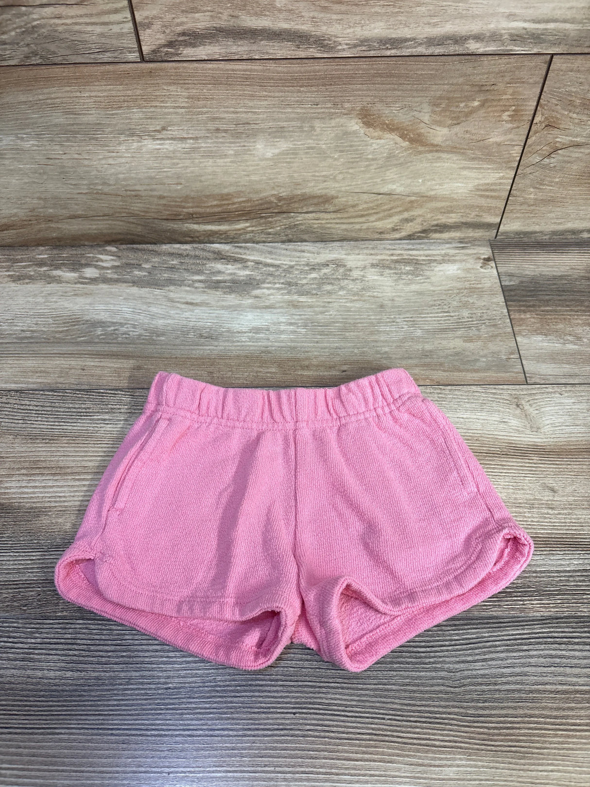 Top-down view of pink terry toddler shorts (2T) showing elastic waistband, side pockets, and curved hem on wood background