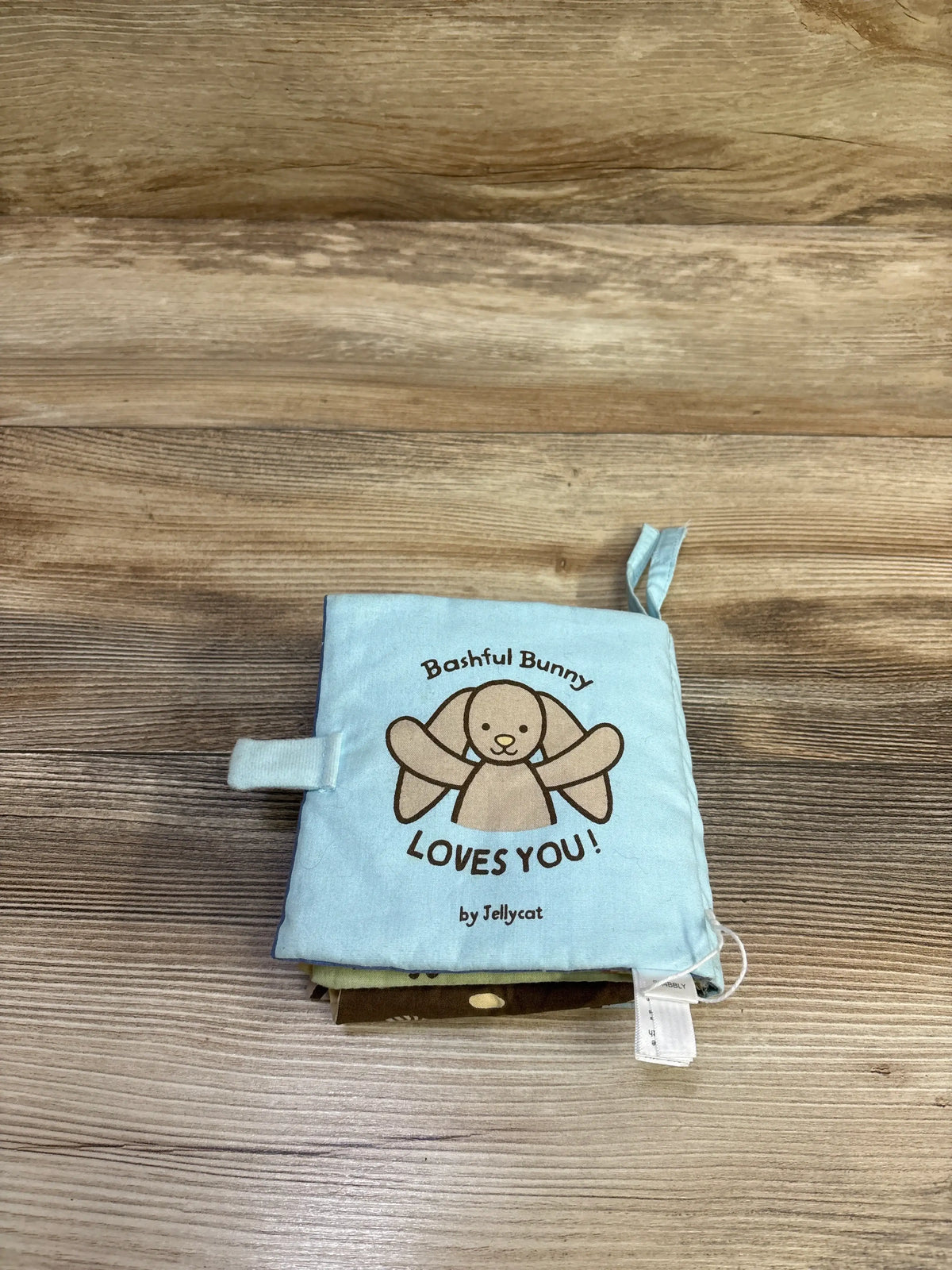 Front view of Jellycat Bashful Bunny Loves You Soft Book showcasing a blue fabric cover with bunny illustration on wood background