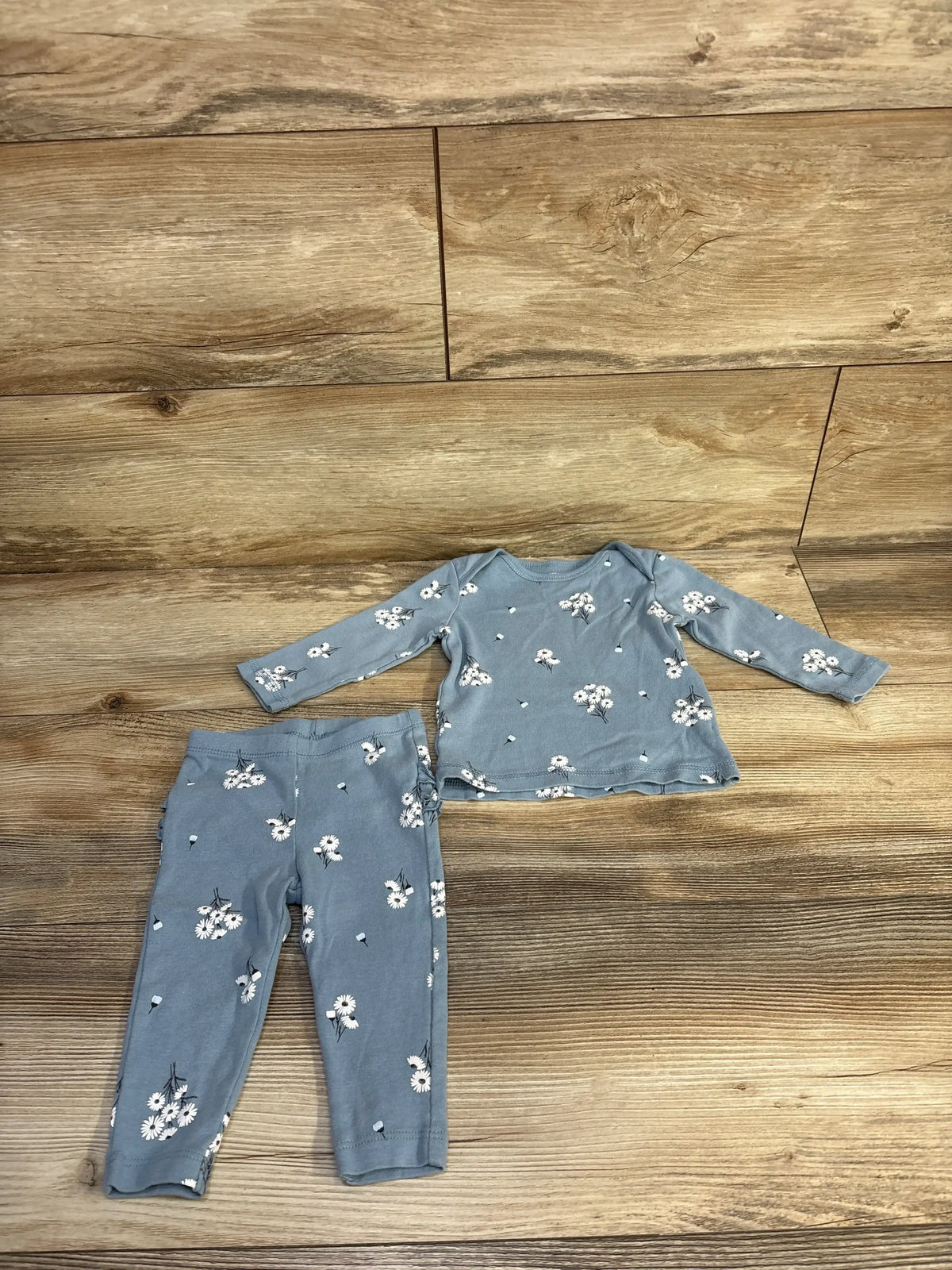 Flat lay of blue Carter’s 2pc shirt and leggings set size 6M showing daisy print, ruffle hem and elastic waist on wood