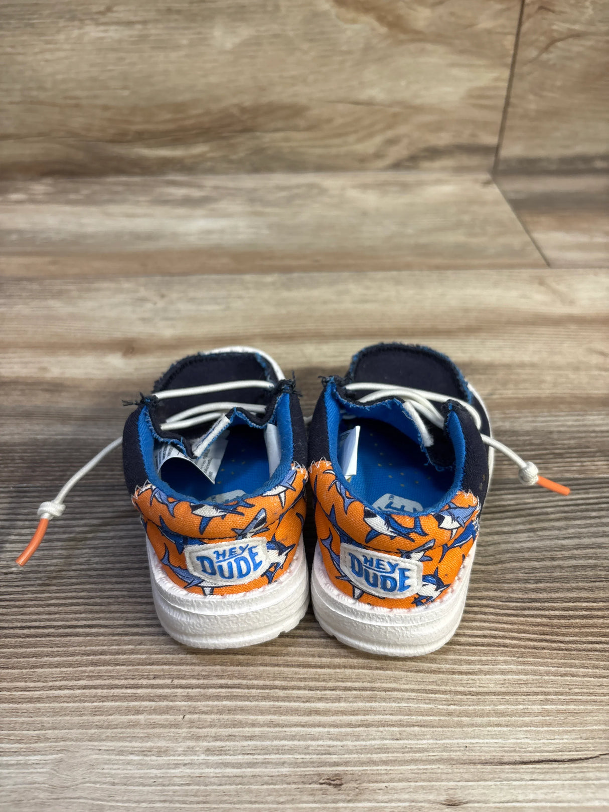 Rear view of Hey Dude Wally toddler shoes showing orange shark-print heel, navy upper, white elastic laces on wood background