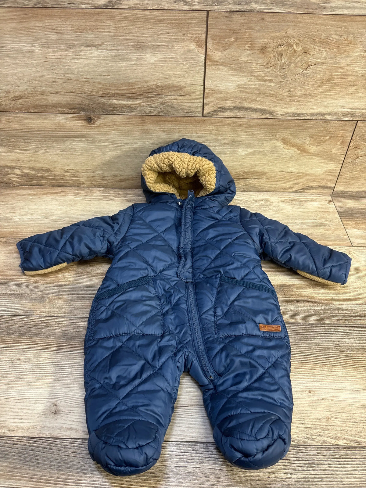 Front view of Perry Ellis navy puffer snowsuit 0–3M, quilted with front zipper and hood lined in tan sherpa on wood floor
