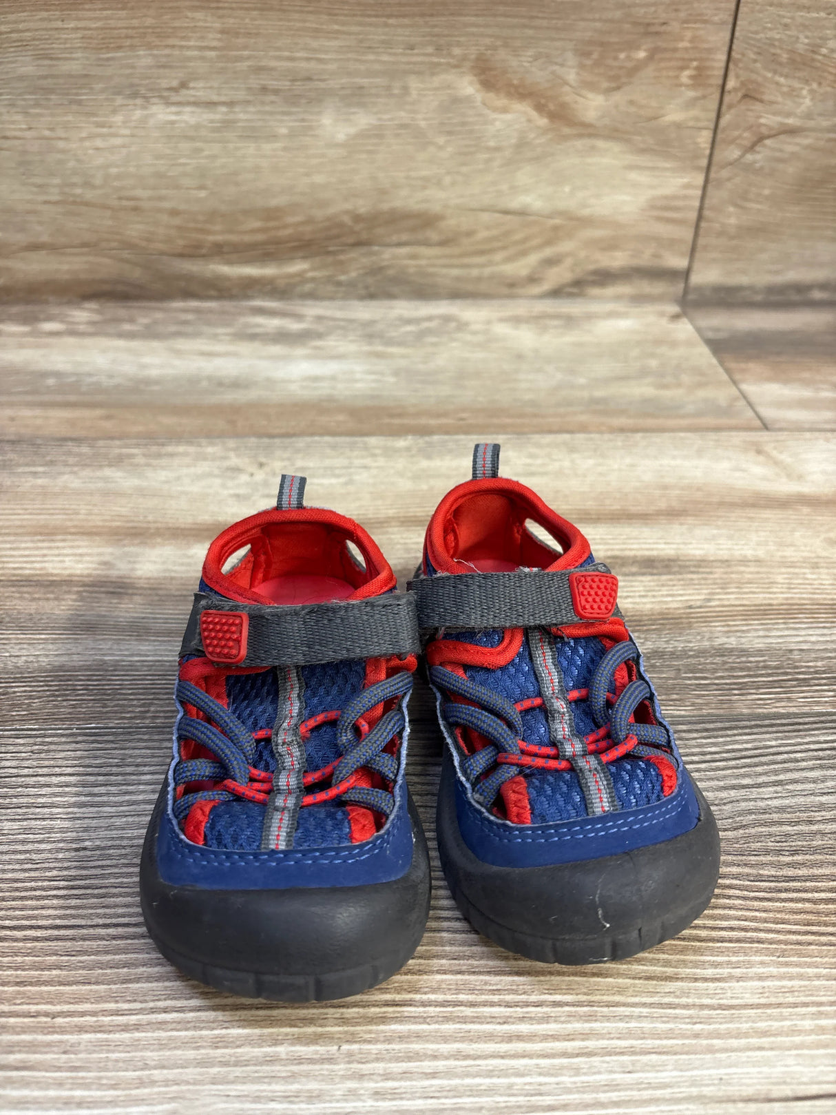 Front view of Oshkosh Pumba2 Bumptoe sandal in navy and red with closed rubber toe, adjustable strap, mesh upper on wood