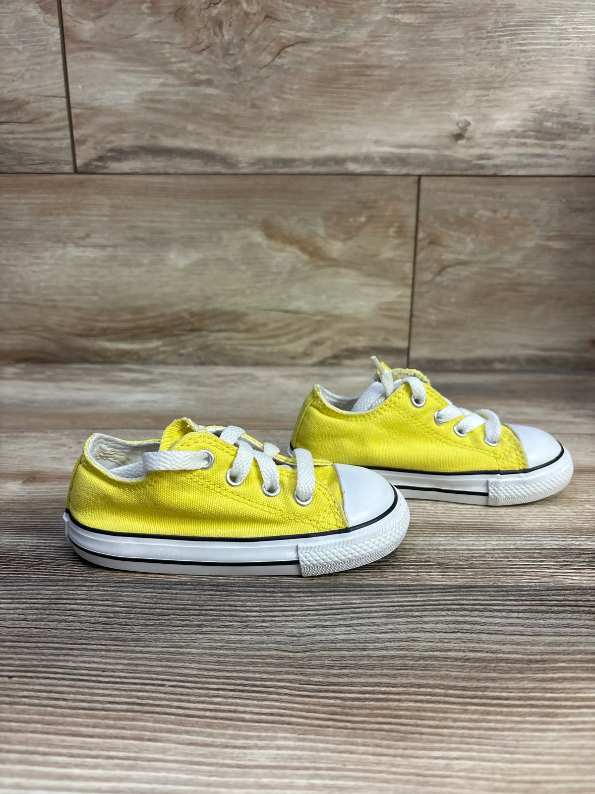 Side view of yellow Converse Chuck Taylor All Star low-top sneakers showing canvas upper, white rubber toe cap and white laces on wood background