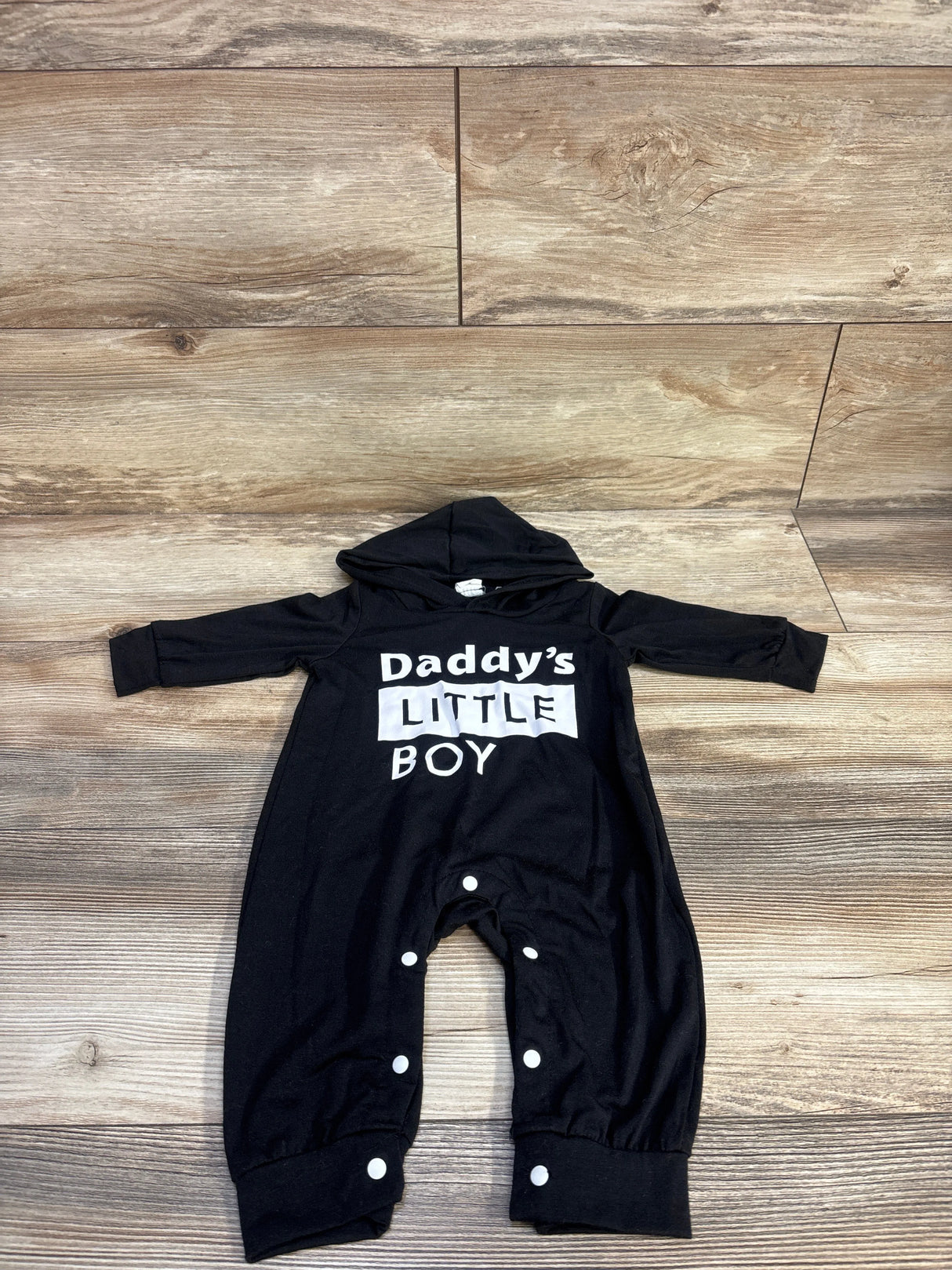 Flat lay of black baby coverall (3–6m) showing white Daddy’s LITTLE BOY print and snap leg closures on wood