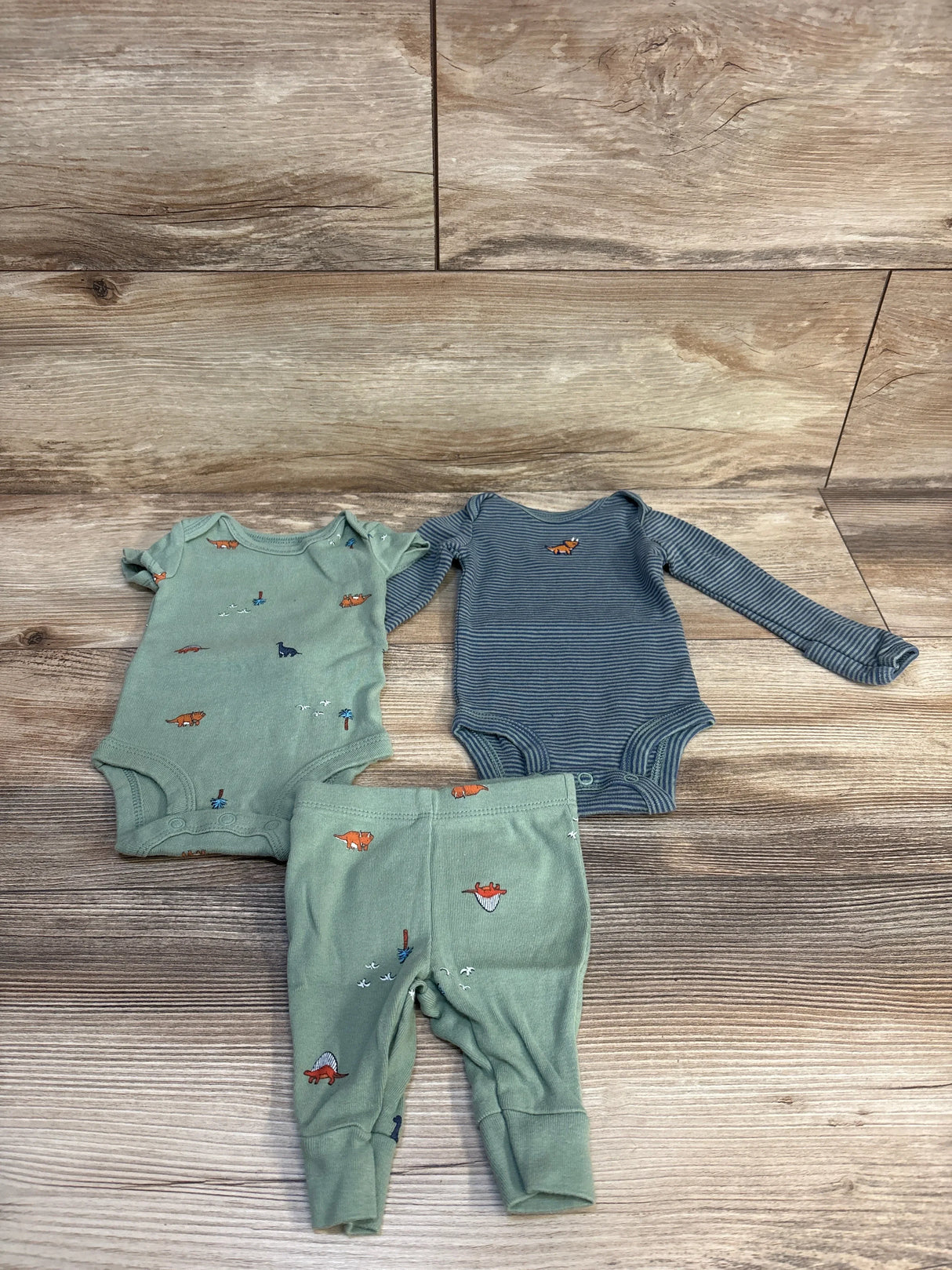 Flat lay of Carter's 3-piece newborn set: green animal-print bodysuit, matching pants, and blue striped bodysuit on wood