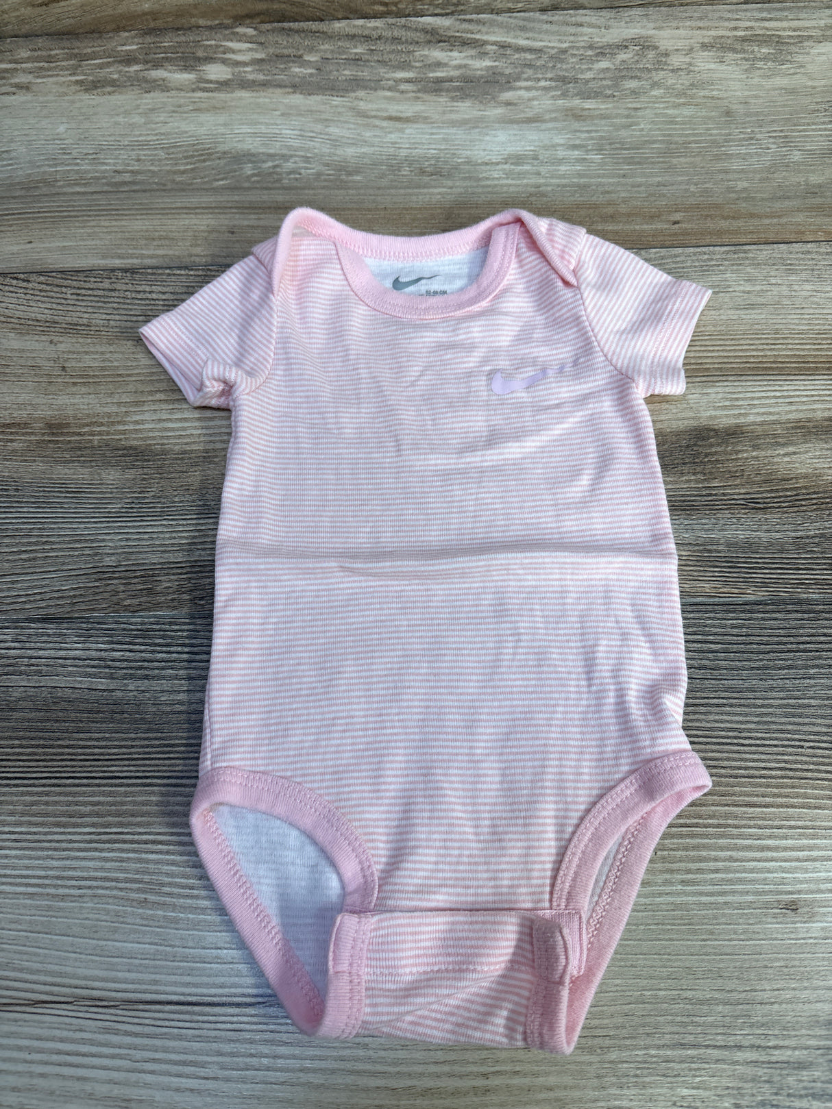 Front view of Nike pink striped bodysuit in size 6m showcasing short sleeves and bottom snaps on a wood-grain background