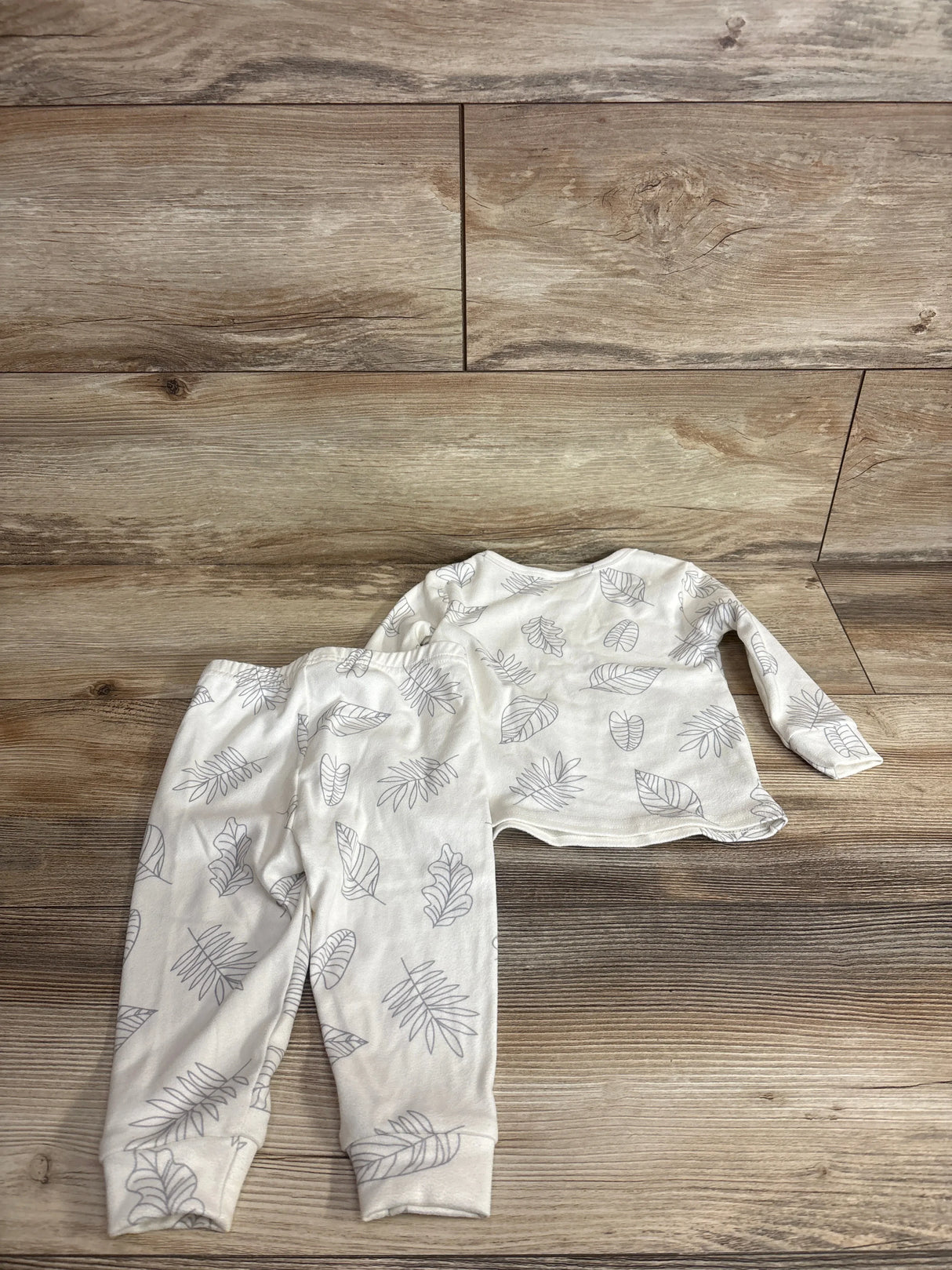 Flat lay of white Cozy Pixies 2-piece baby pajama set size 9 to 12 months showing gray leaf print on wood floor background
