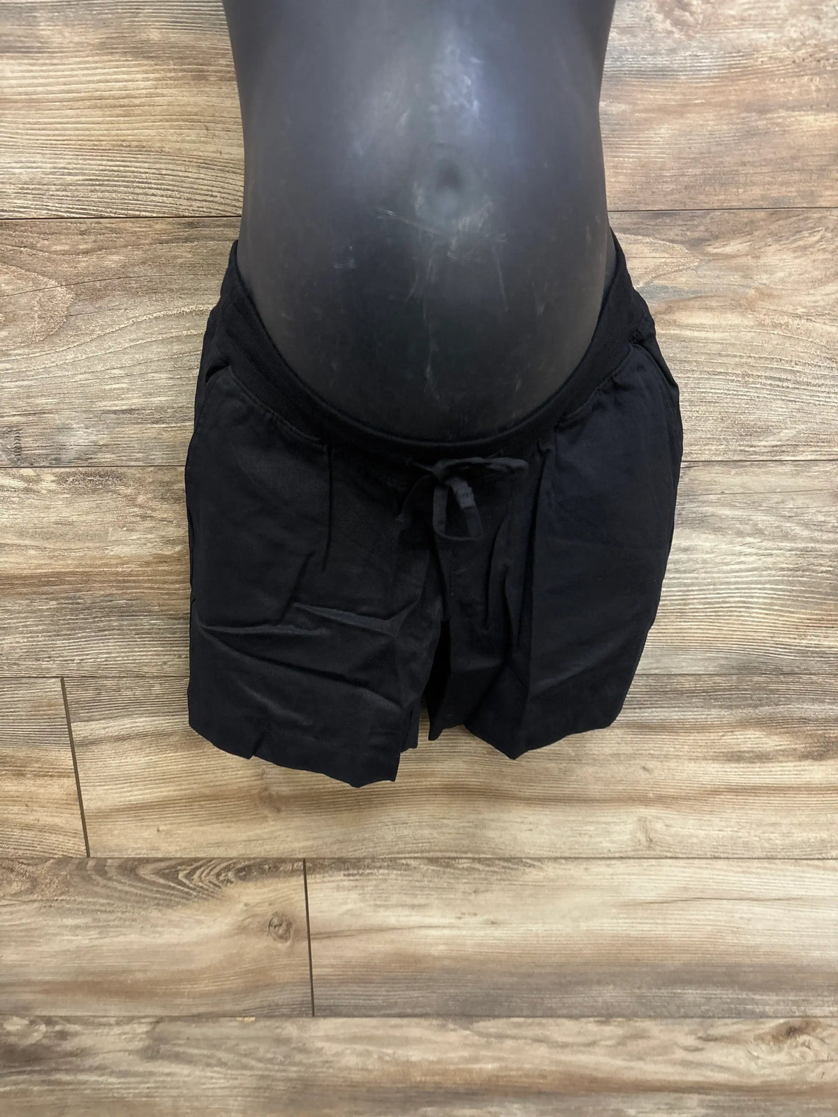 Front view of black Gap maternity linen-blend shorts showing under-belly waist, drawcords and slant pockets on wood-look floor