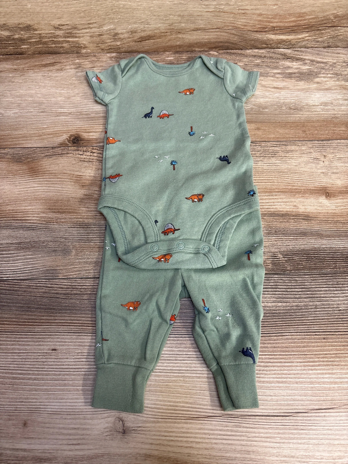 Flat-lay of green Carter's 2-piece baby bodysuit and pants showcasing orange and navy dinosaur print, snap crotch, and ribbed ankle cuffs on a wood background