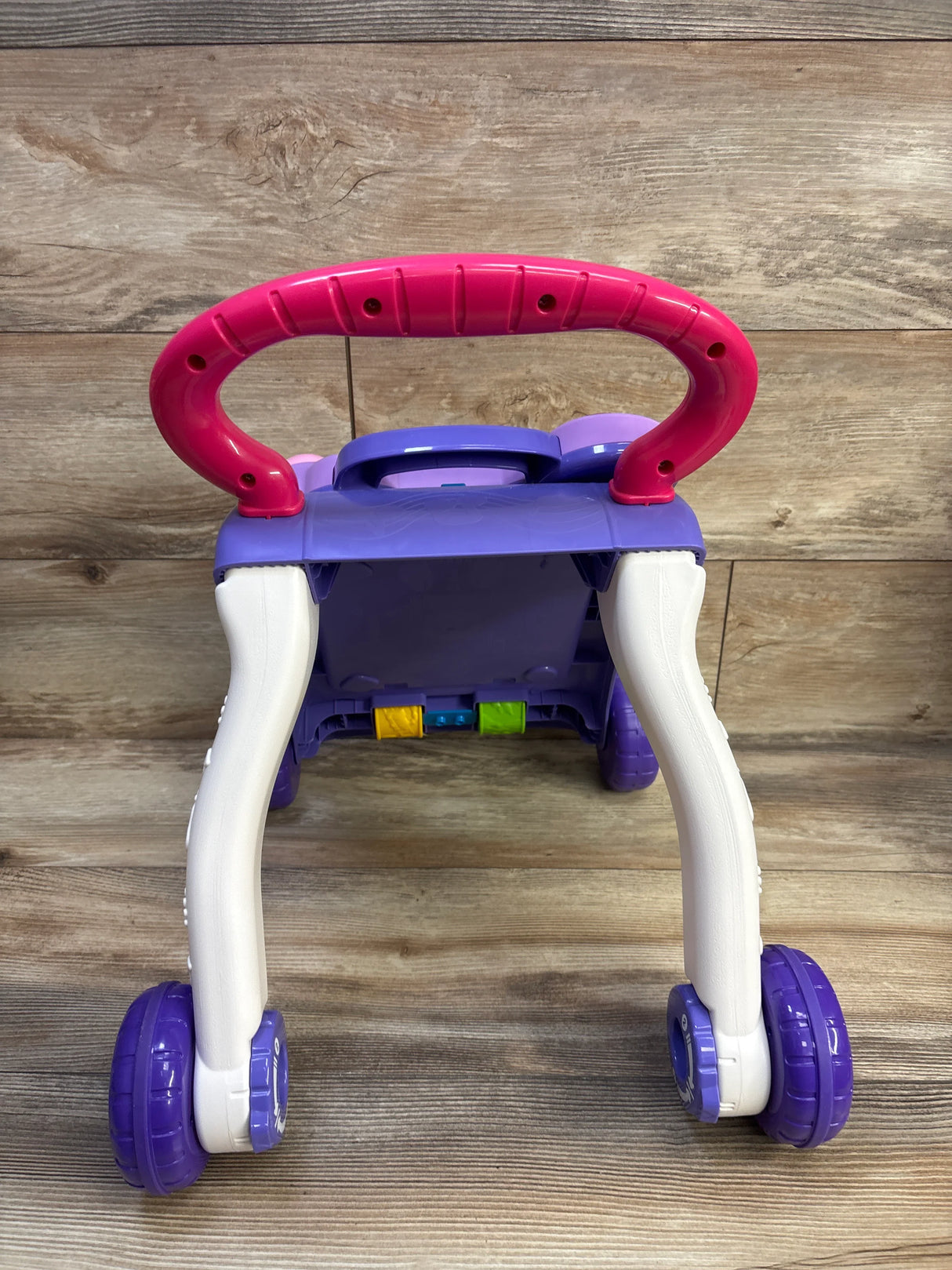 Rear view of VTech Sit-to-Stand Walker with pink handle, white legs, purple wheels, and colorful blocks on wood backdrop