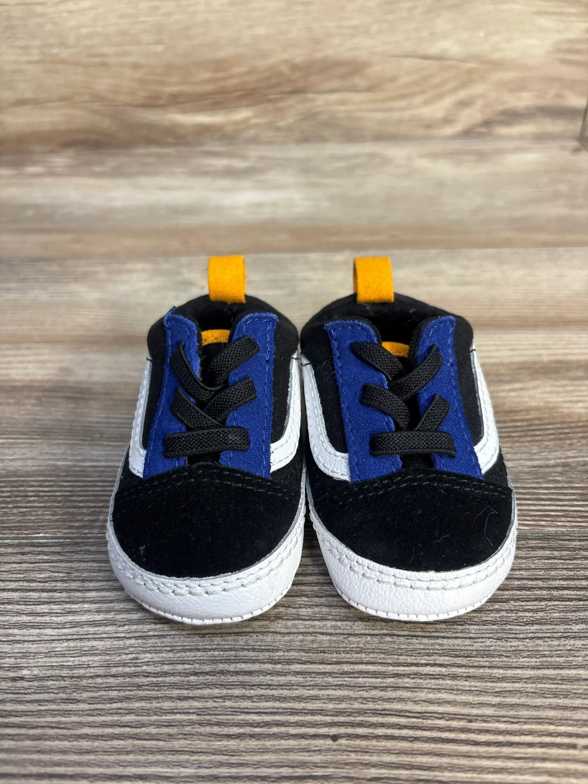 Front view of black Vans Old Skool crib shoes in size 2C with blue tongues, white stripes, and heel pull tabs on wood grain