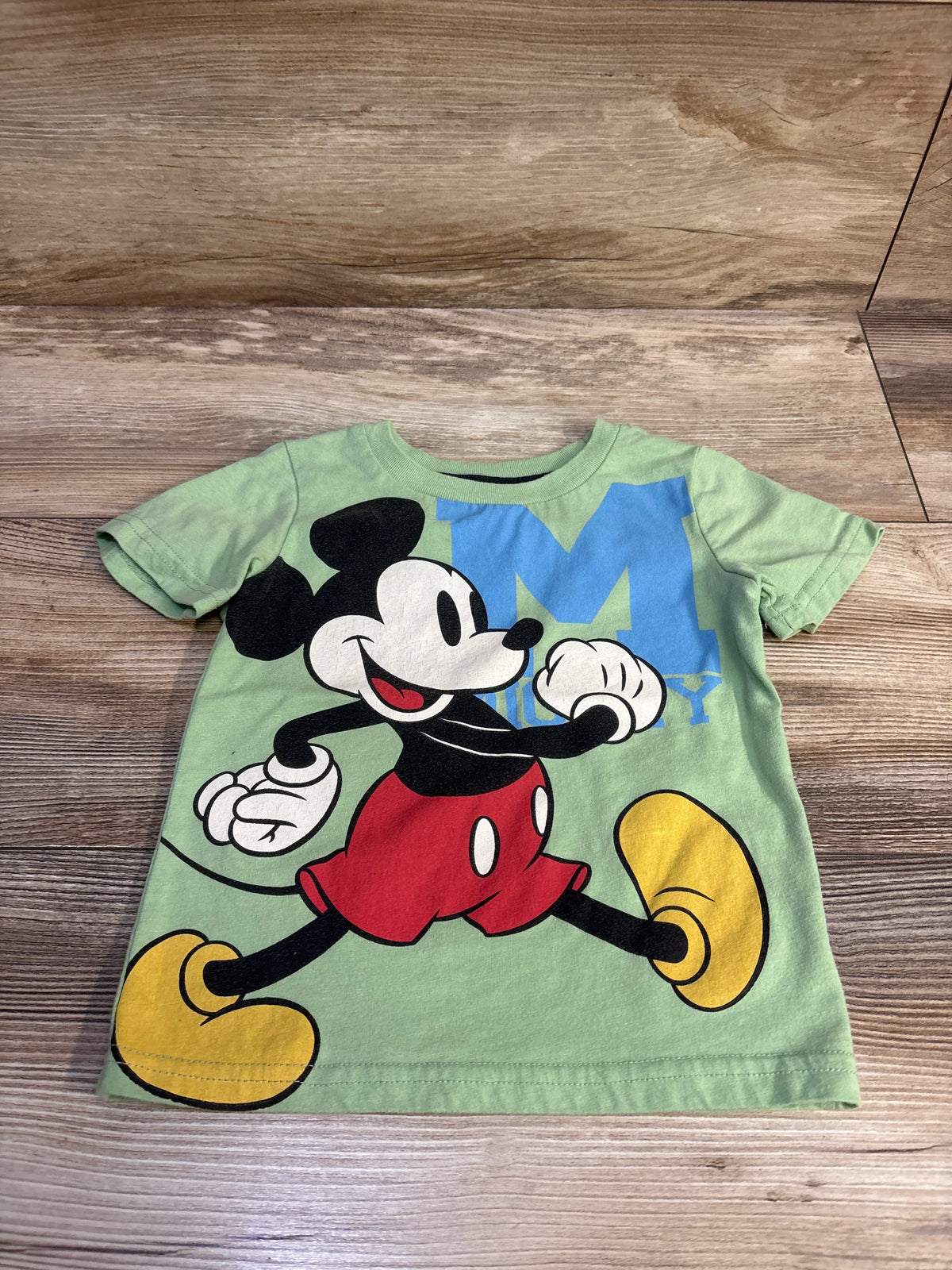 Front flat-lay of green Mickey Mouse toddler t-shirt size 2T showcasing large printed Mickey graphic and blue ‘M’ on wood background