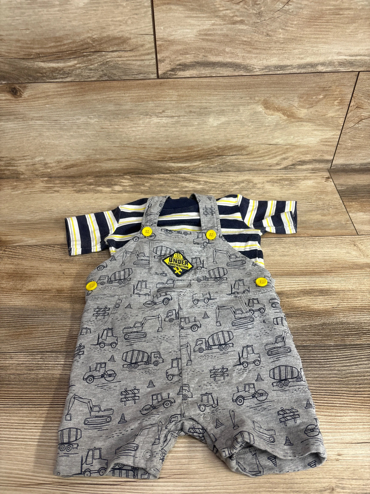 Flat lay of navy-striped baby shirt and gray construction-print shortalls showcasing yellow button accents on wood floor