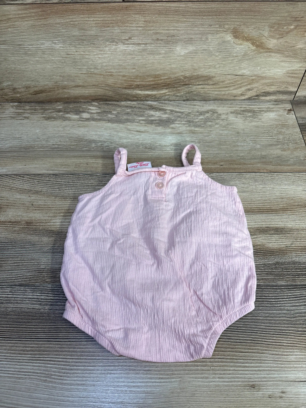 Flat lay of pink Little Lass bubble romper with two buttons, thin straps and elastic leg openings on a wood floor