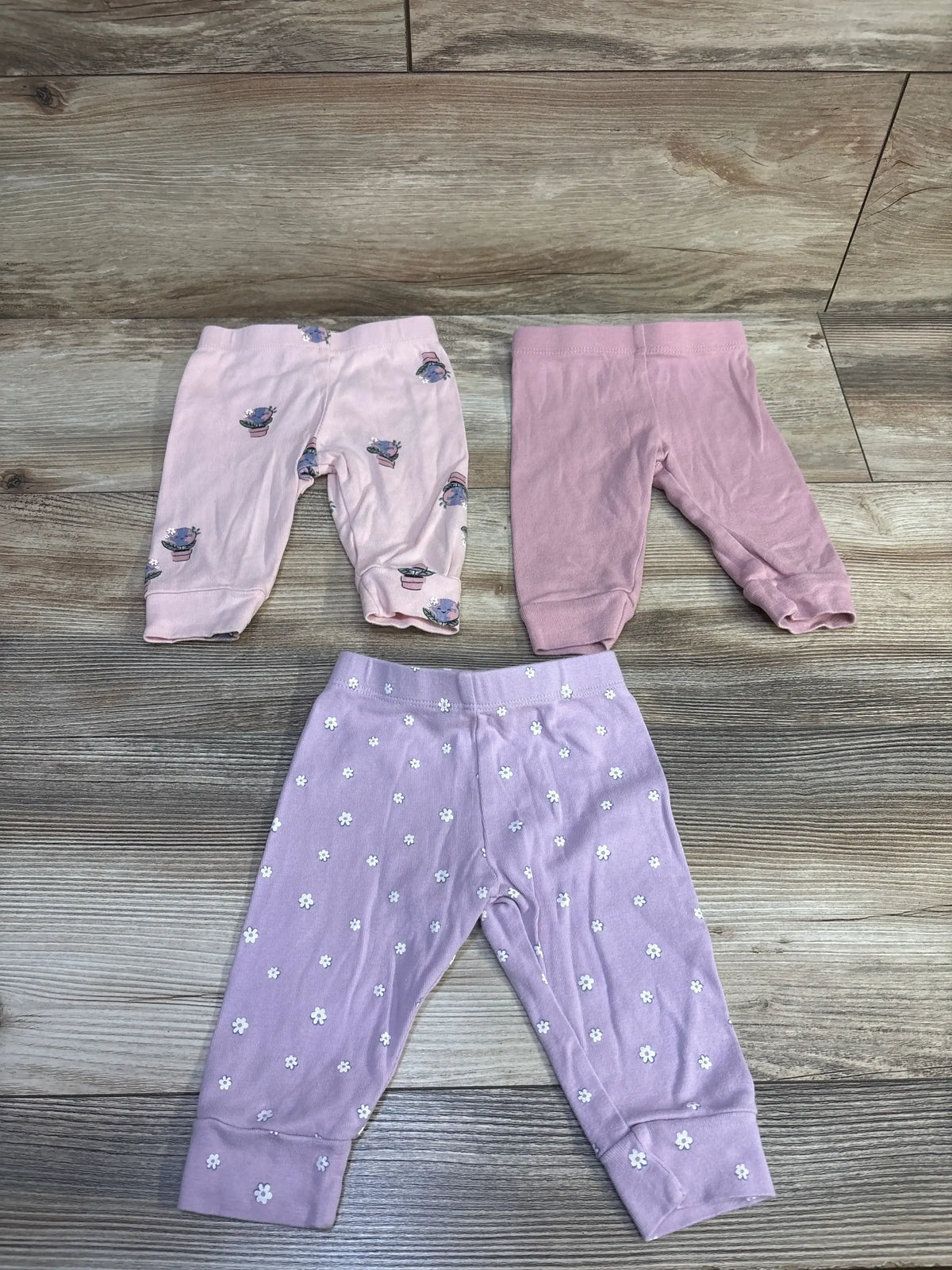 Front view of Pekkle three-pack baby pants showcasing pink printed, solid, and floral designs on a wood-look floor