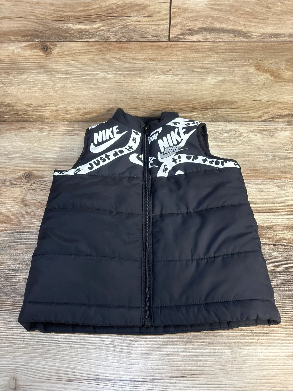 Kids black puffer vest with zipper and white graphic logo print, displayed on wood floor