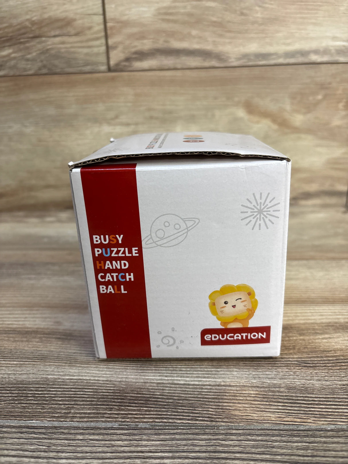 Front view of Happy Play Puzzle Busy Ball box showing red side-panel text and cartoon lion on a wooden surface
