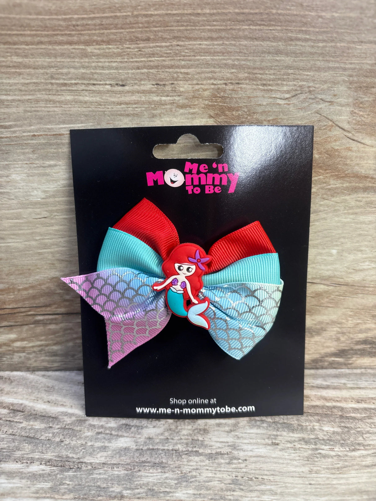 Front of mermaid hair bow showcasing red and teal ribbons, scale-pattern tails, and mermaid center on black display card