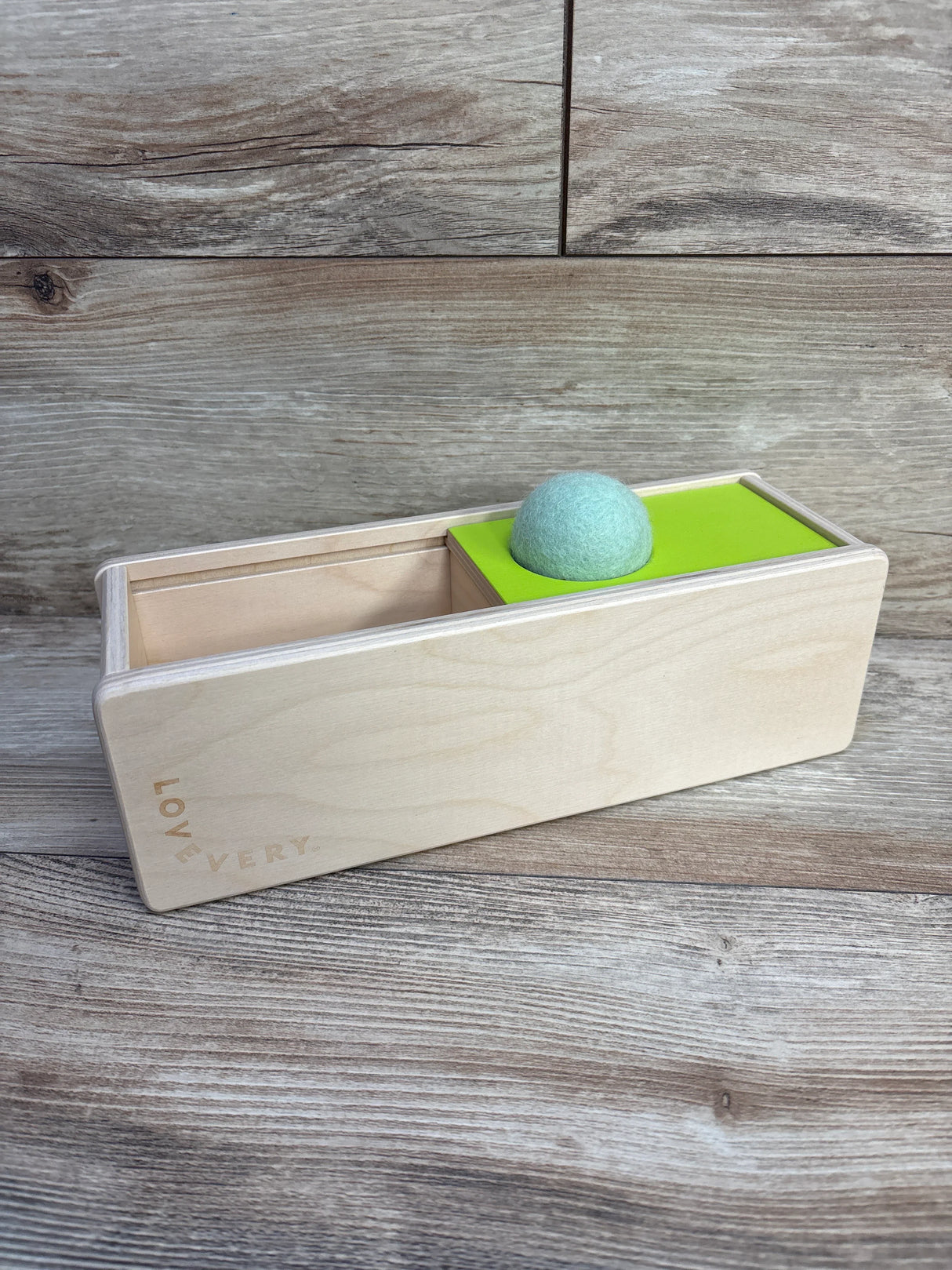Wooden baby toy with sliding box and green felt ball by Lovevery on wood surface