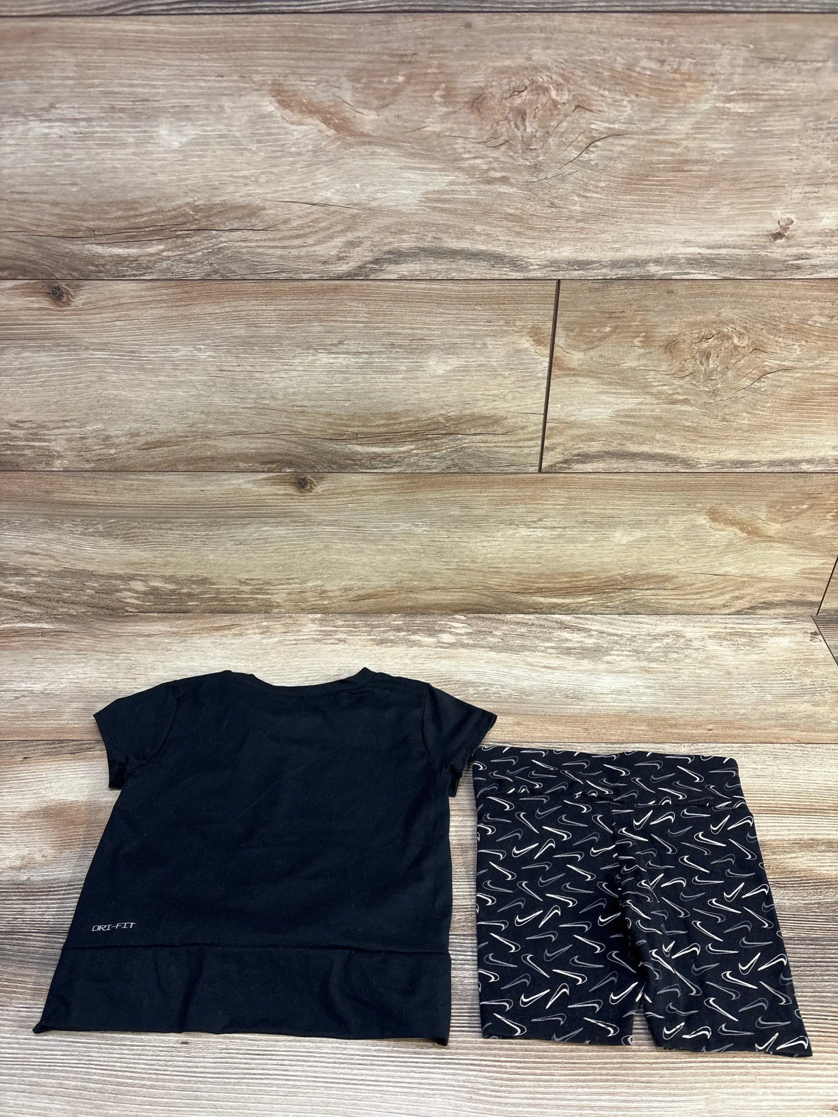 Flat lay of Nike Dri-Fit black shirt and matching shorts size 4T with Dri-Fit hem logo and white swoosh print on wood floor