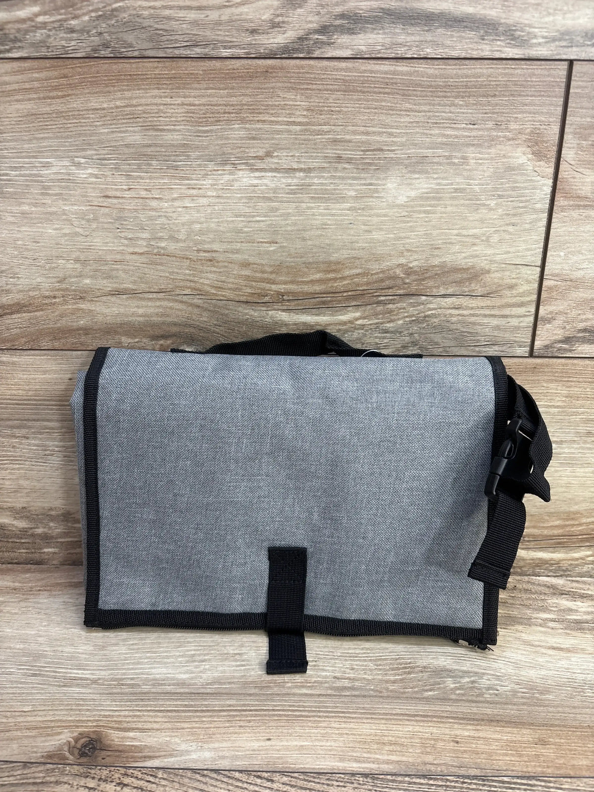 Front view of the gray backpack changing station showcasing its stroller attachment strap and closure strap on a wood surface