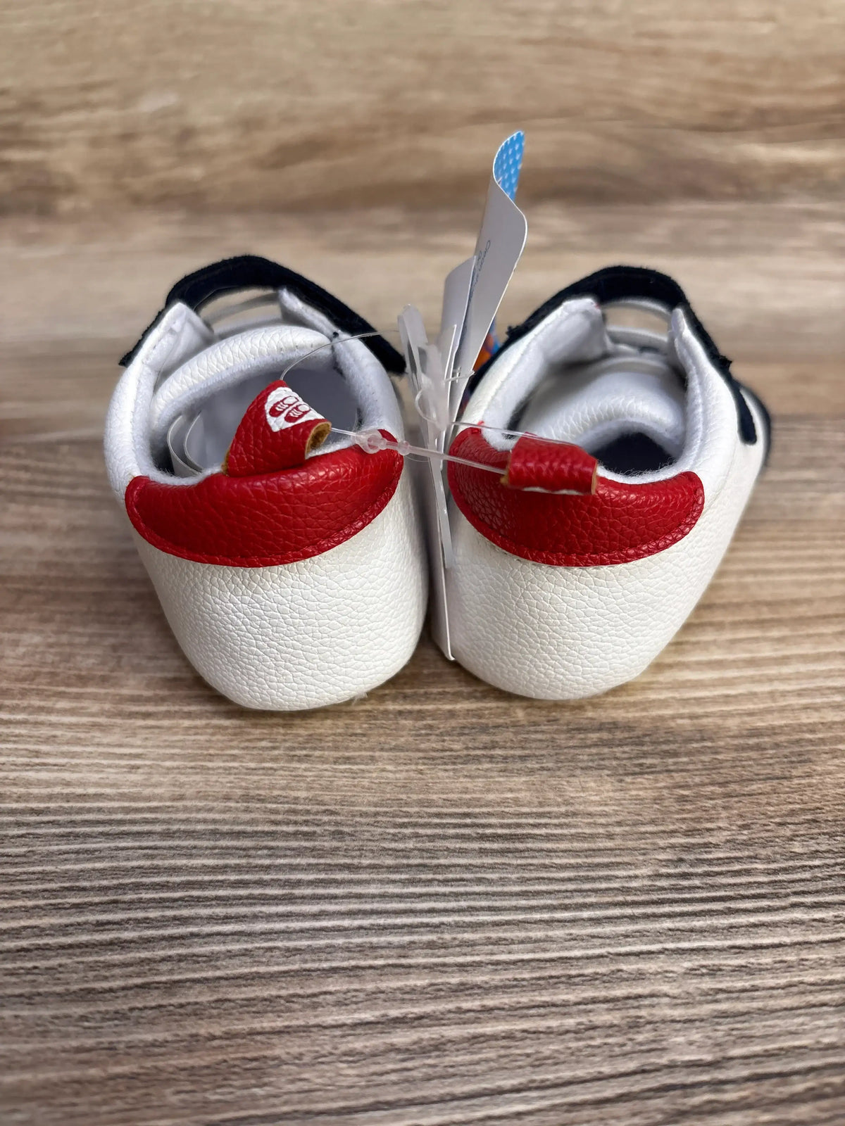 Rear view of pair of white baby baseball shoes showing red heel accents and navy straps on wood surface