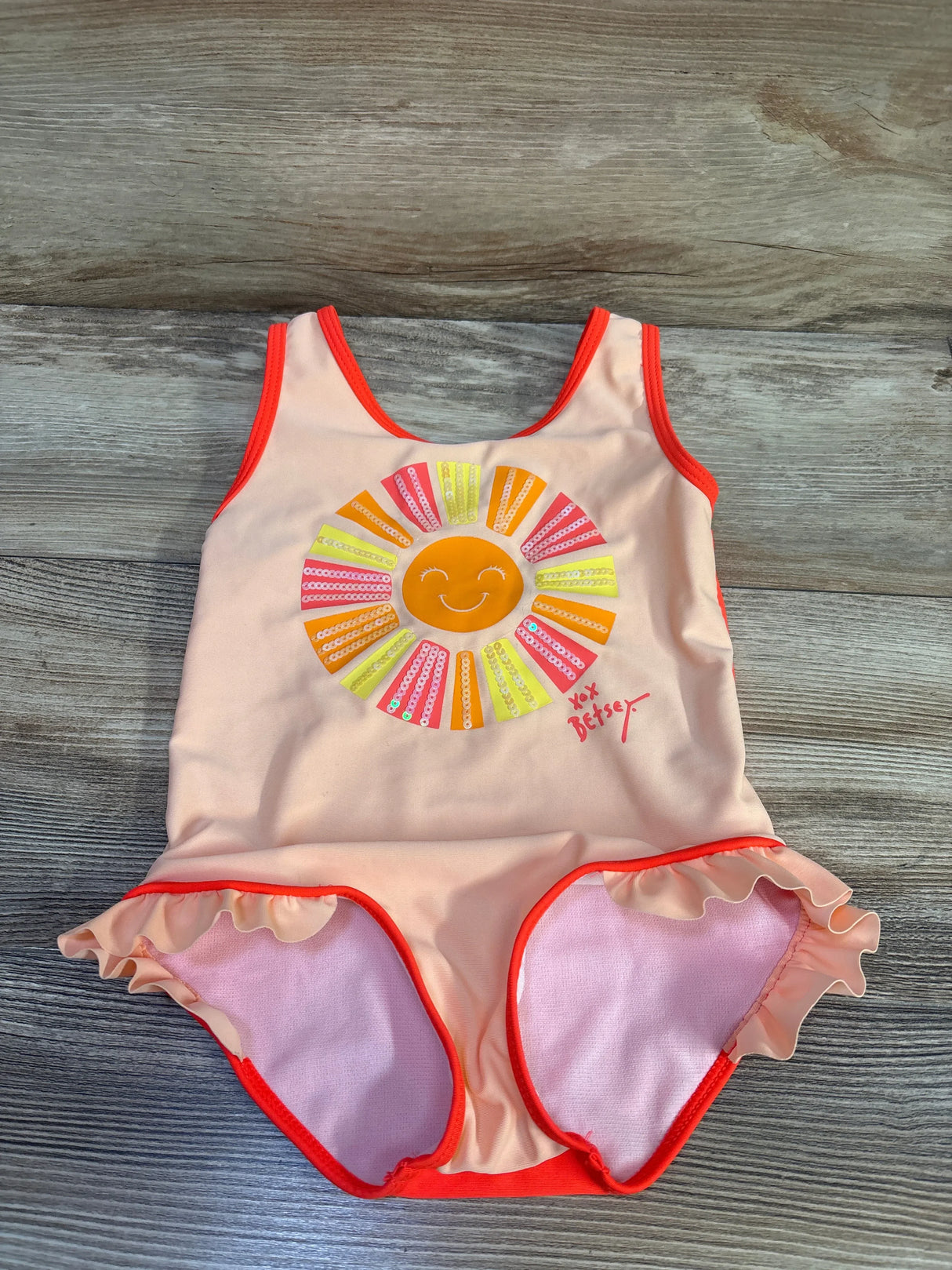 Front flat-lay of girls one-piece swimsuit with smiling sequin sun, ruffled leg trim and bright orange binding on wood background