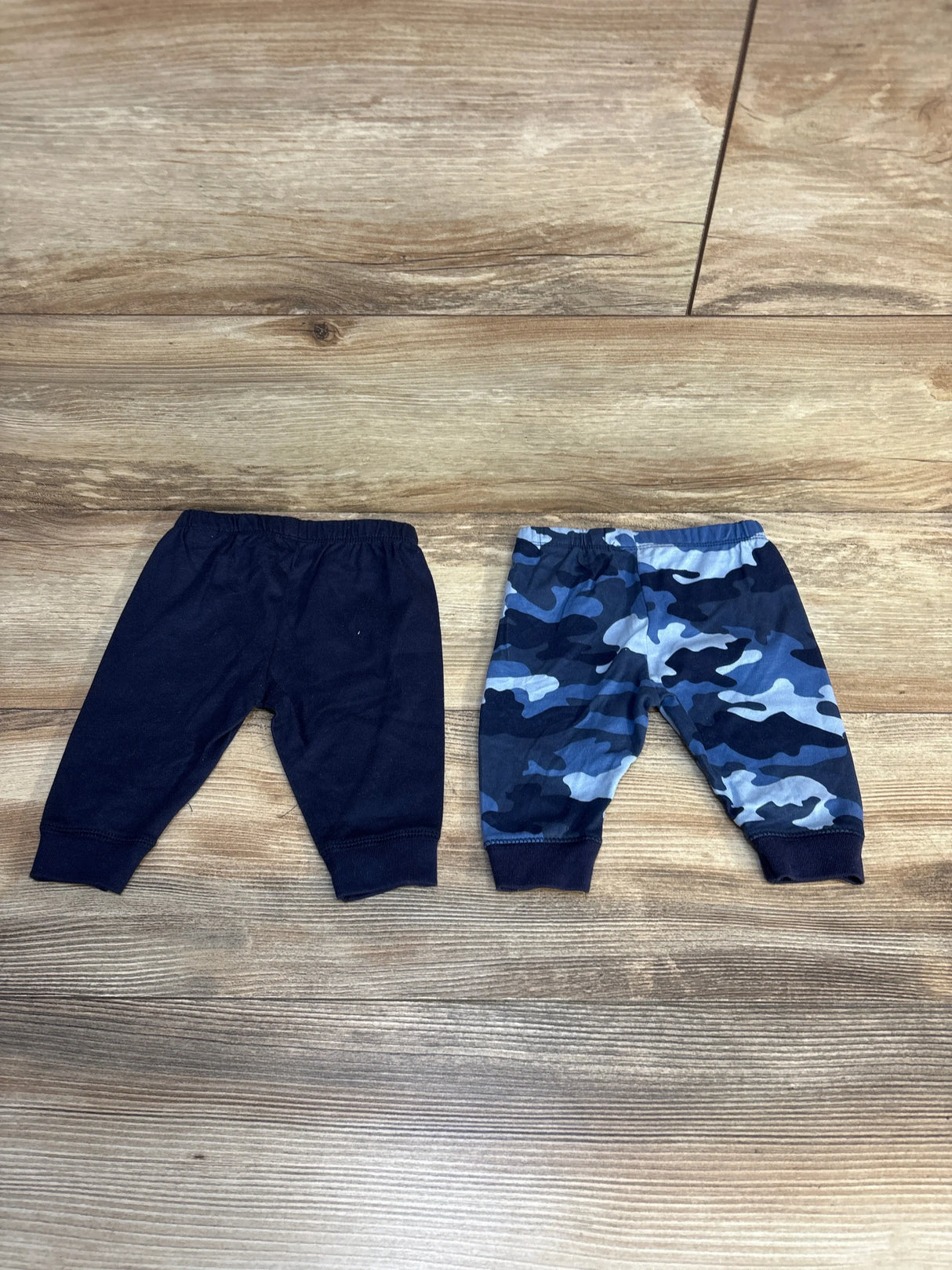 Flat lay of baby pull-on pants 2pk showcasing solid navy and blue camo prints with elastic waistbands on wood plank floor