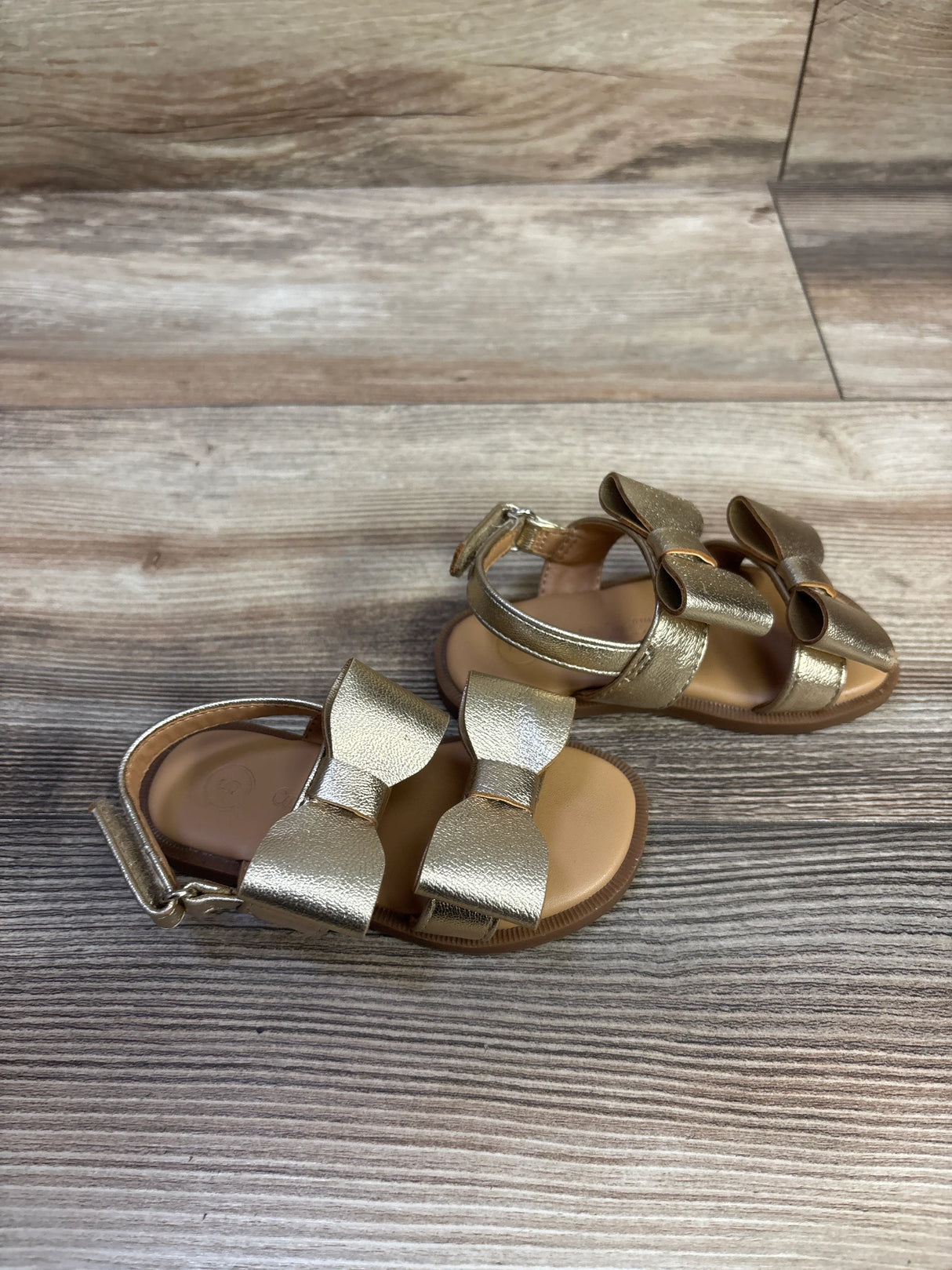 Angled top view of Cat & Jack Ruby bow sandals showcasing metallic gold finish, double bows, and ankle strap on wood-grain background