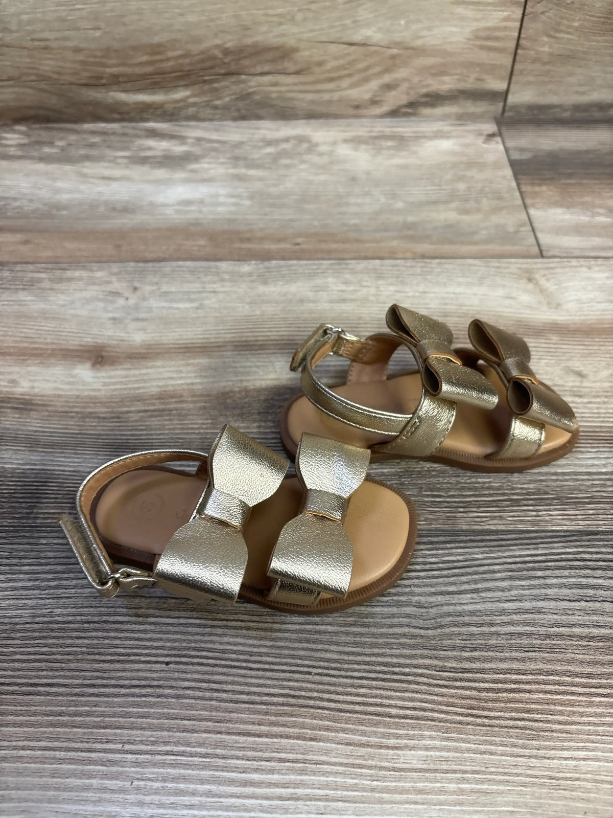 Angled top view of Cat & Jack Ruby bow sandals showcasing metallic gold finish, double bows, and ankle strap on wood-grain background