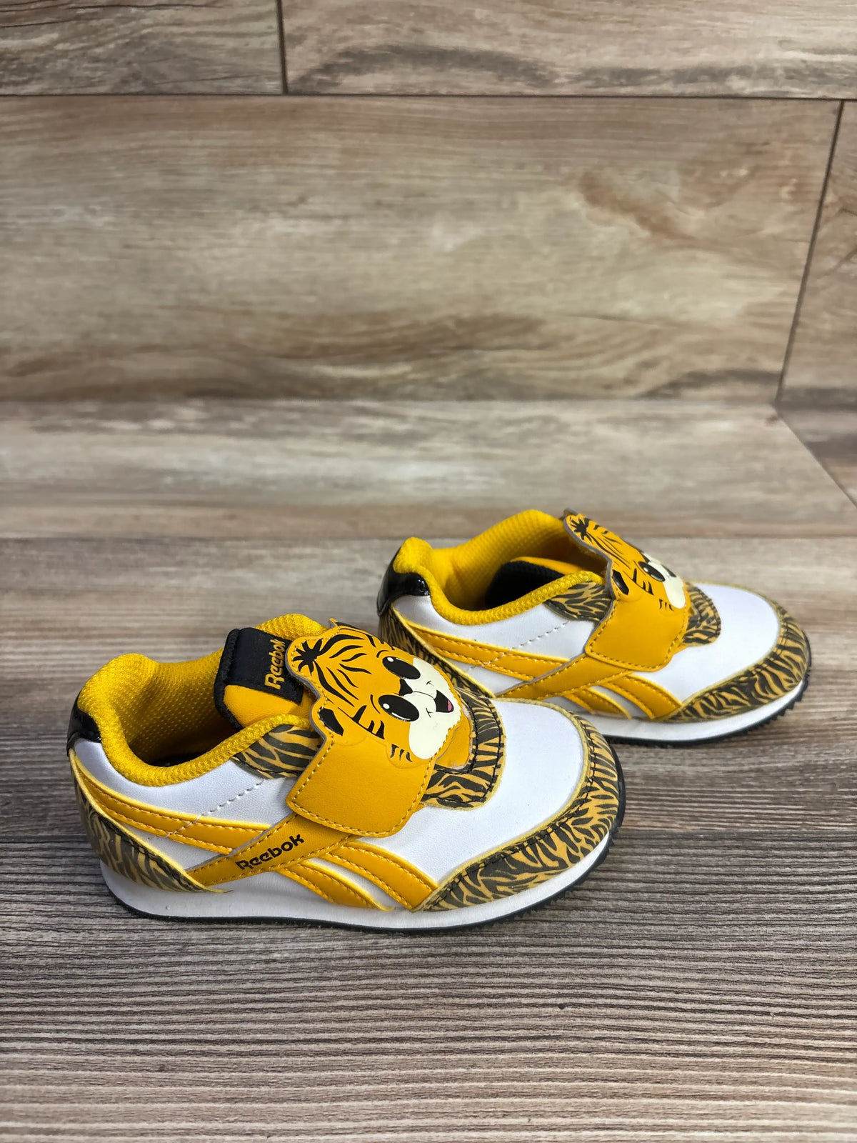 Three-quarter view of Reebok Royal Classic 2.0 sneakers, size 7C, with tiger graphics and yellow straps on wood floor